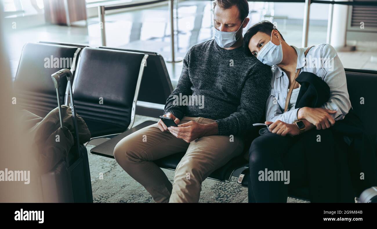 Gelangweilte Touristen, die während einer Pandemie am Flughafen gestrandet sind. Mann, der am Flughafen neben einer Frau mit dem Handy sitzt. Beide in Gesichtsmasken. Stockfoto