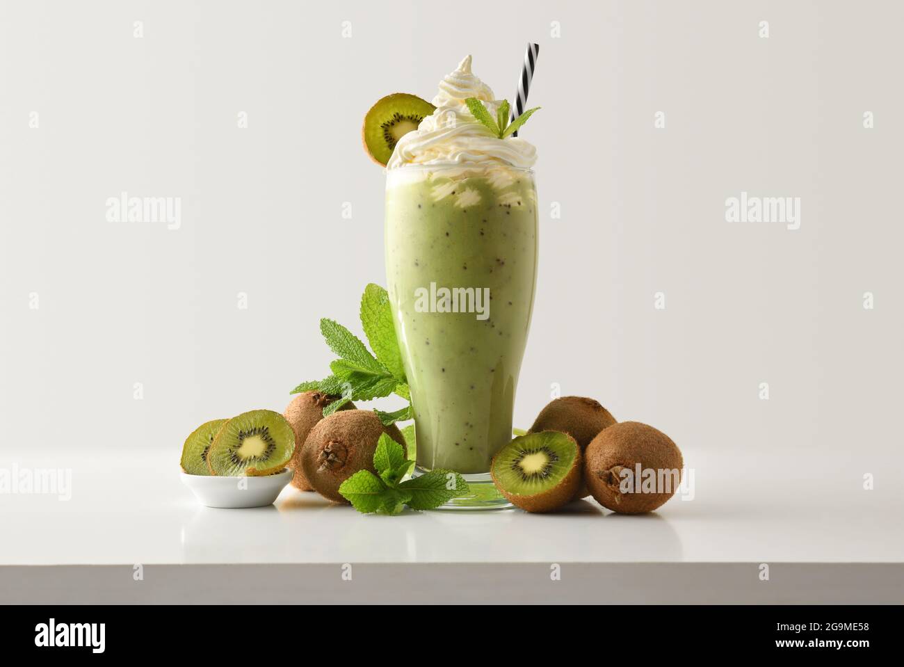 Kiwi-Milchshake mit cremefarbenem Dekor, verziert mit Früchten und Blättern auf weißem Tisch und hellem, isoliertem Hintergrund. Vorderansicht. Stockfoto