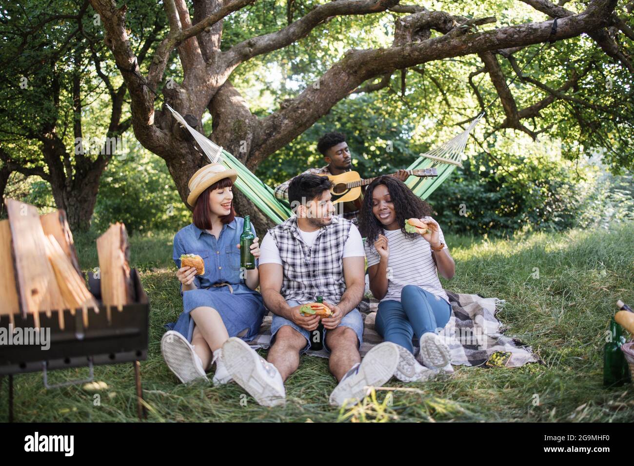 Drei junge Leute, die im Sommergarten auf Plaid sitzen und leckere Burger essen und Bier trinken. Verwischen Sie den Hintergrund eines afrikanischen Mannes, der in der Hängematte Gitarre spielt. Picknick von diversen Freunden. Stockfoto