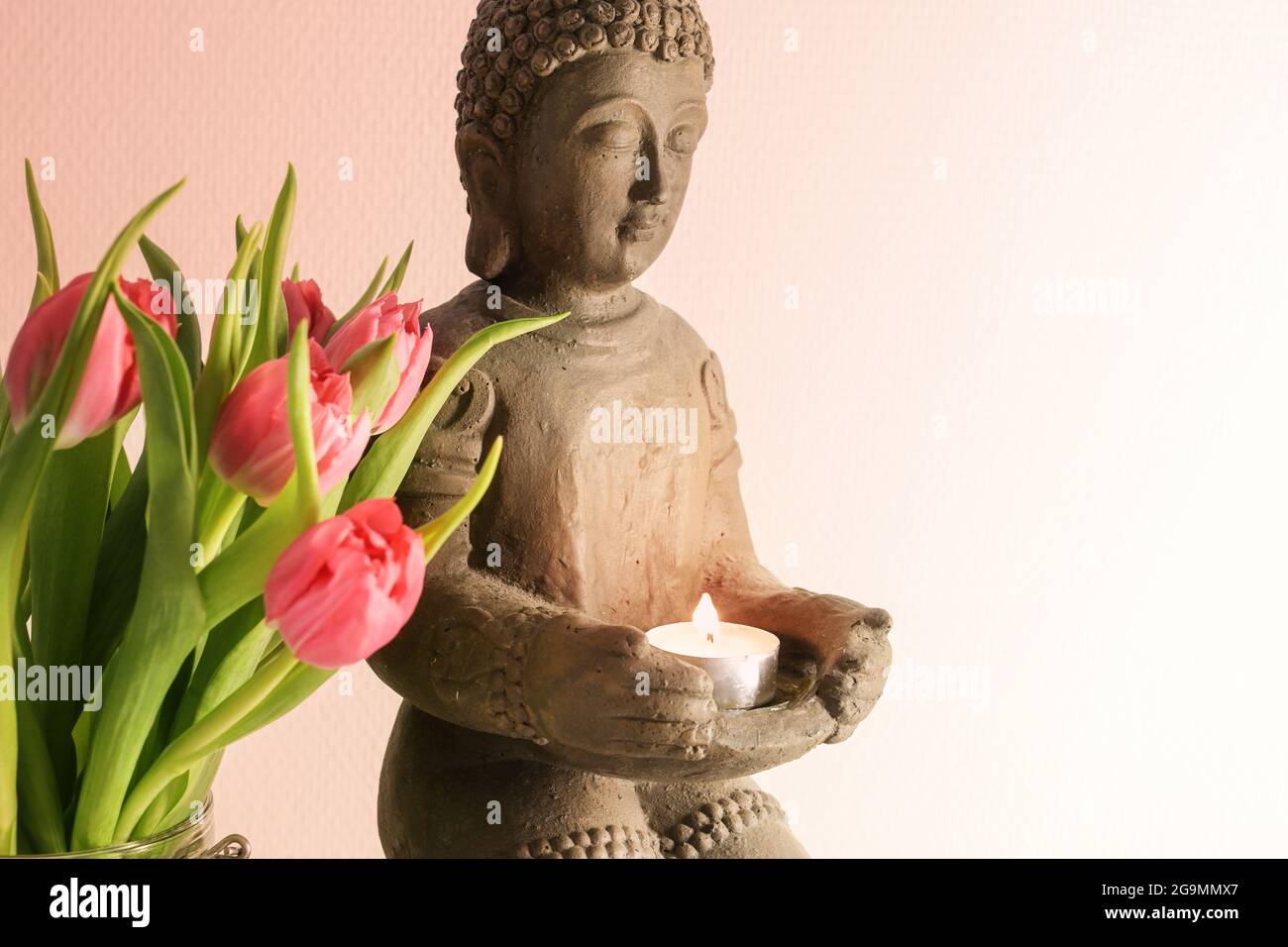 Kleine buddha-Skulptur aus Keramik mit brennender Kerze und rosa Tulpen vor hellem Hintergrund mit Kopierraum, ausgesuchter Weichfokus, schmal Stockfoto