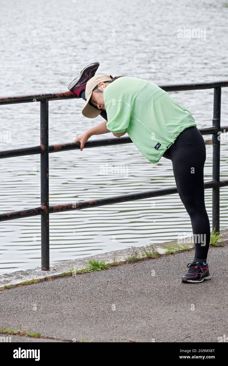 Eine sehr flexible Frau streckt ihren Oberschenkel auf einen Zaun in der Nähe des Sees im Kissena Park, Flushing, Queens, New York City. Stockfoto