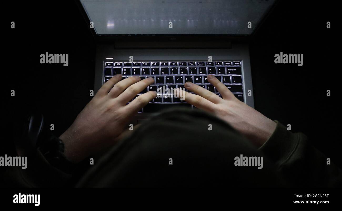 Ein einfaches, sehr dunkles Nachtbild mit Händen auf einer beleuchteten Tastatur, die tippt. Schattige Person, die im Dunkeln an einem Computer oder Laptop eine Kapuze trägt. Stockfoto