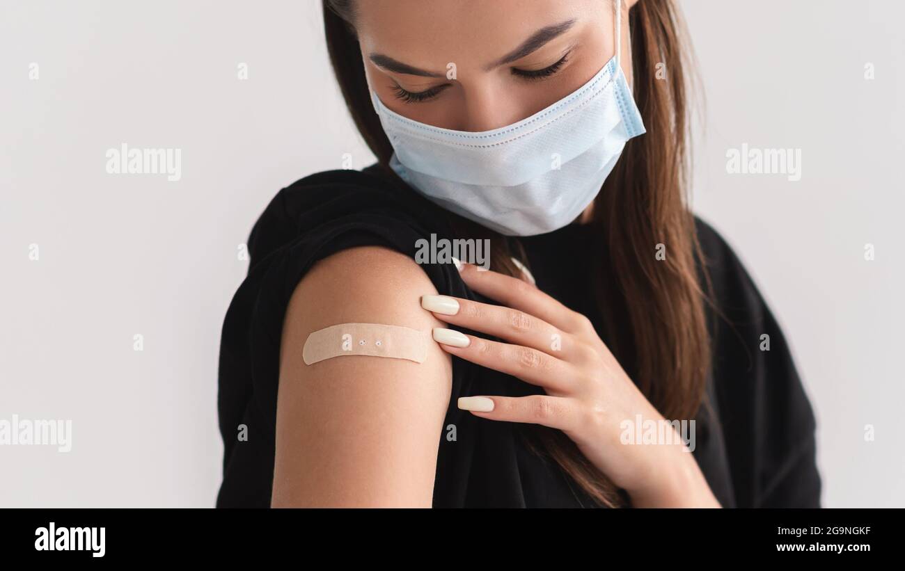 Junge Frau in Gesichtsmaske, die den Arm mit Bandhilfe nach Injektion des Coronavirus-Impfstoffs über hellem Studiohintergrund zeigt Stockfoto