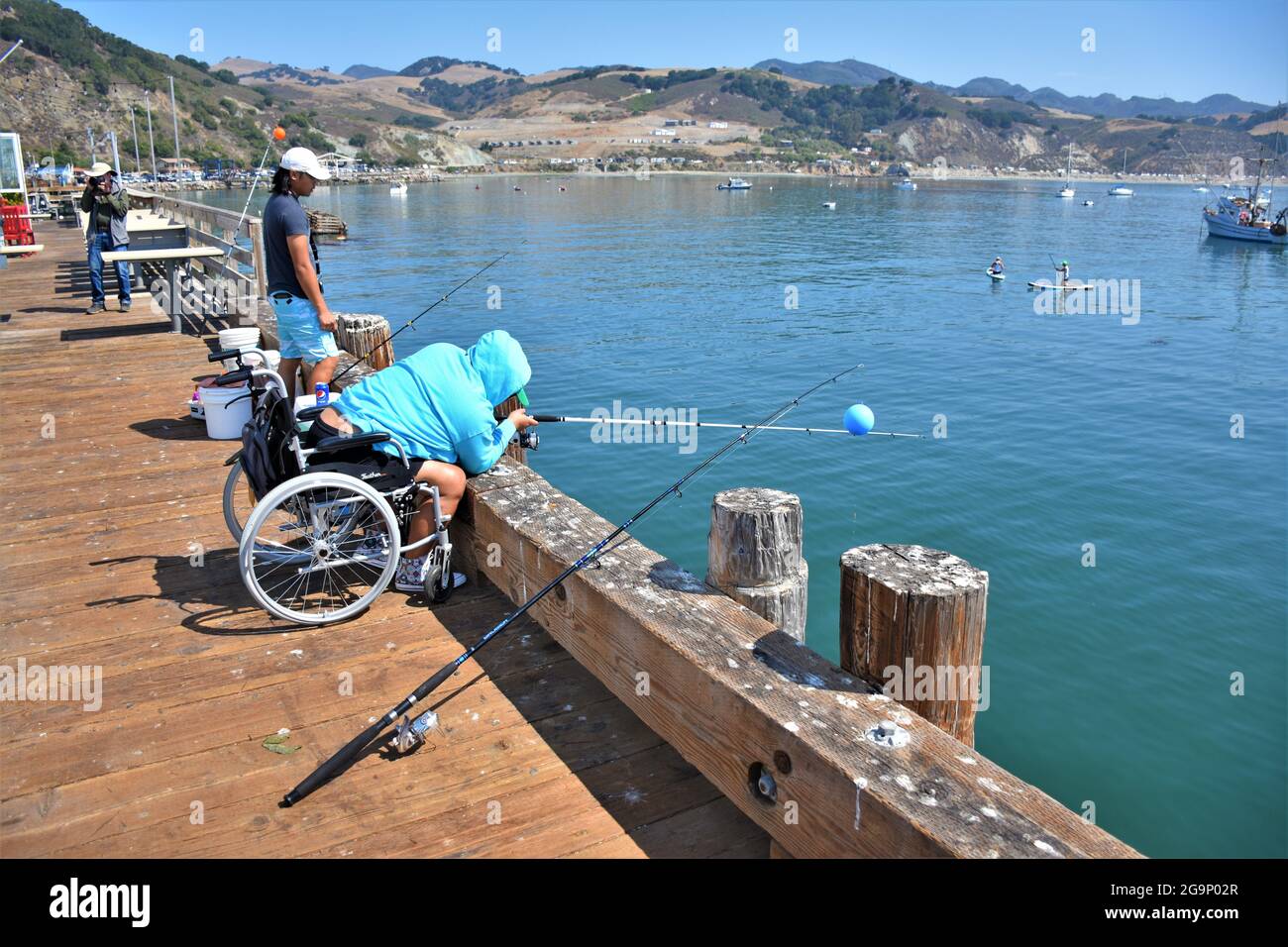 Frau im Rollstuhl, die am öffentlichen Pier in Port San Luis, Avila, Kalifornien, mit Kapuzenpullover angeln muss, an sonnigen Tagen am Strand mit der Familie Stockfoto