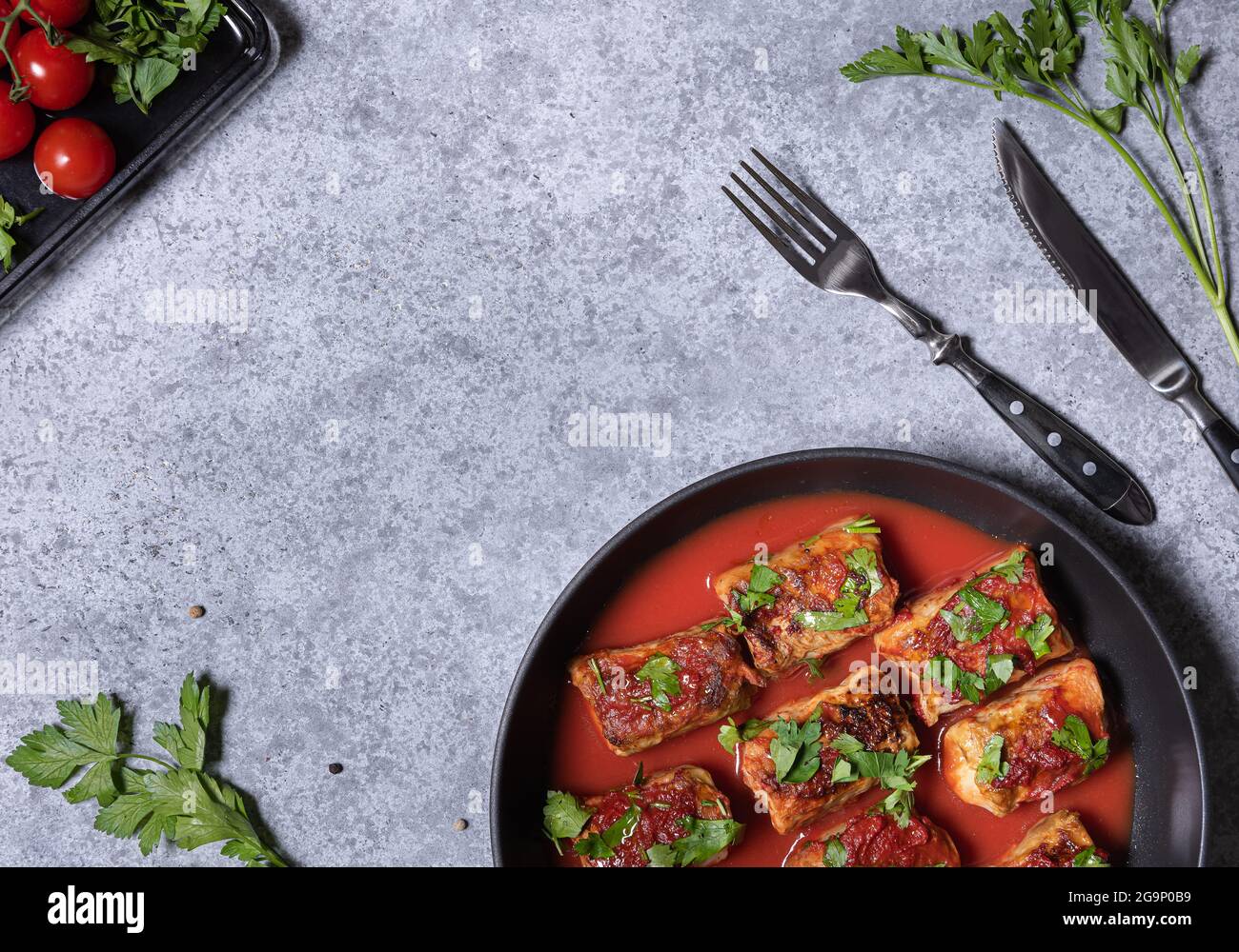 Kohlröllchen, Tomatensauce und frisch gehackte Petersilie in der Pfanne. Flach mit Kohlrollen, Tomaten, Petersilie, Gabel und Messer auf grauer Struktur legen Stockfoto
