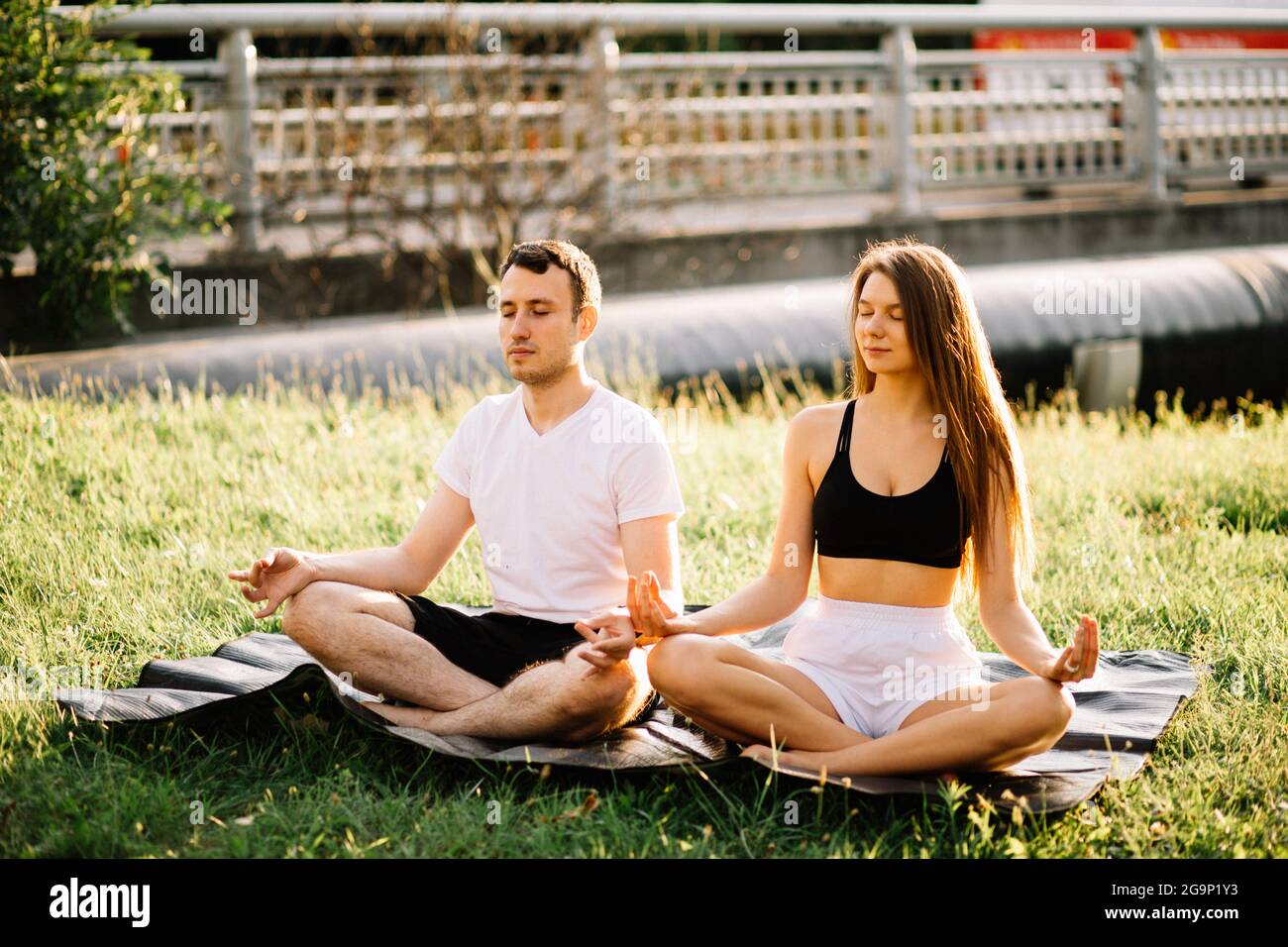 Junge Paar Mann und Frau meditieren, während sie in Lotusposition sitzen, Yoga auf dem Rasen der Stadt, Sommerabend Zweisamkeit Stockfoto
