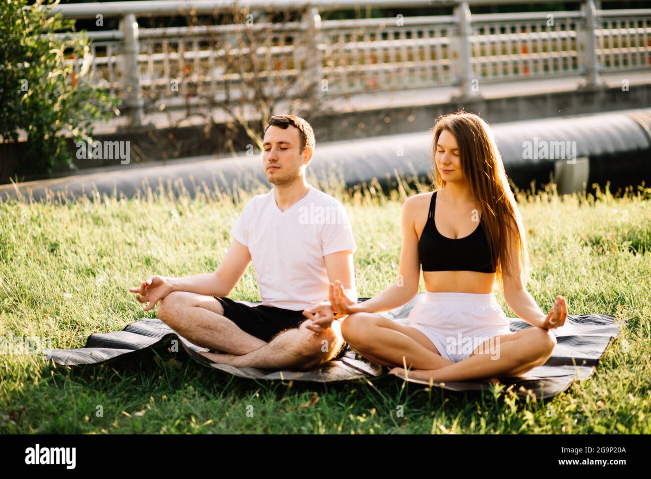 Junge Paare, Mann und Frau, meditieren in Lotusposition, Yoga auf dem Rasen der Stadt, Sommerabend zusammen Stockfoto