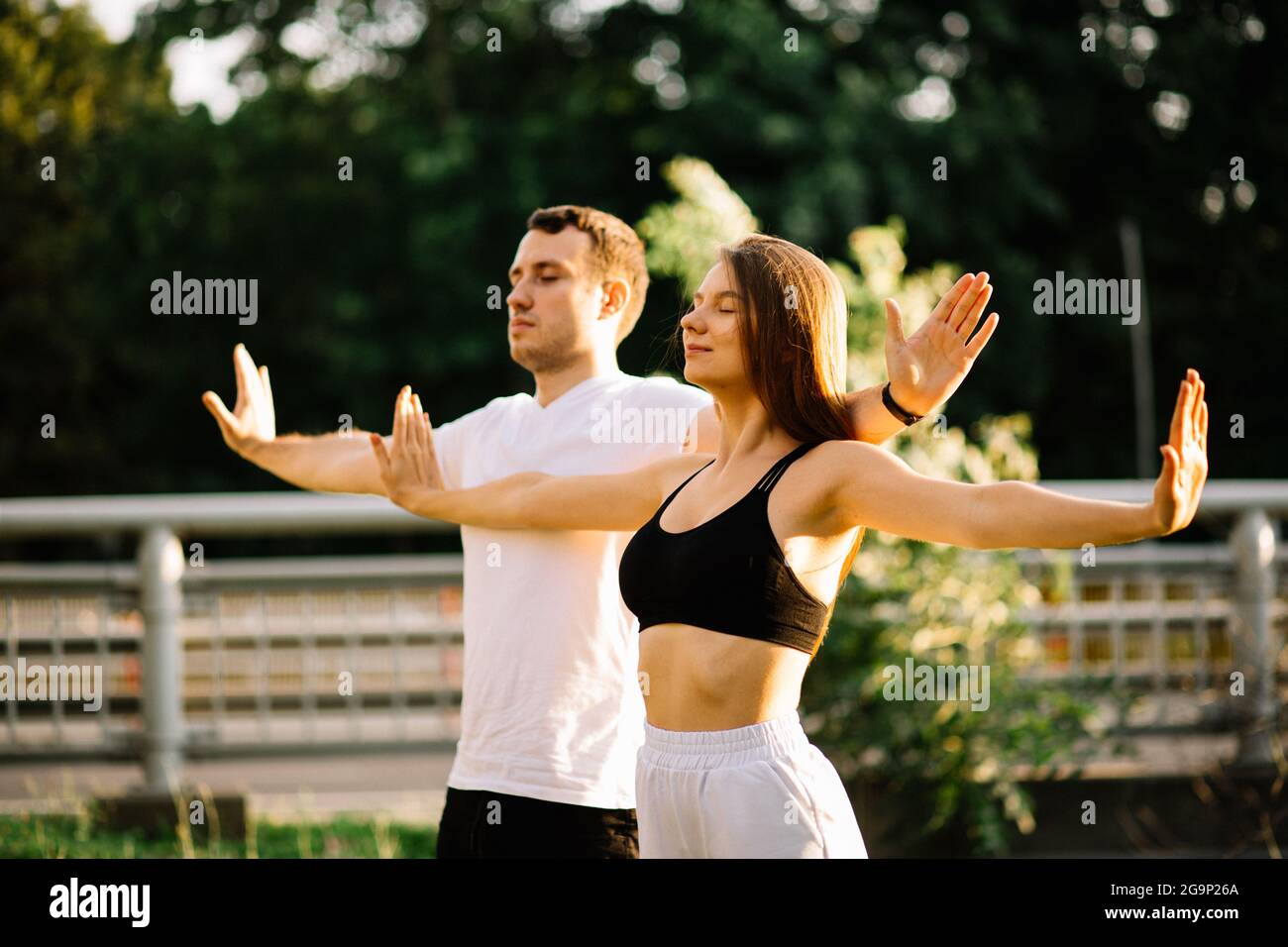 Das junge Paar, Mann und Frau, meditieren gemeinsam beim Sonnenuntergang, während sie stehen, Yoga auf dem Rasen, Sommerabend, Stadtleben, Atemübungen Stockfoto