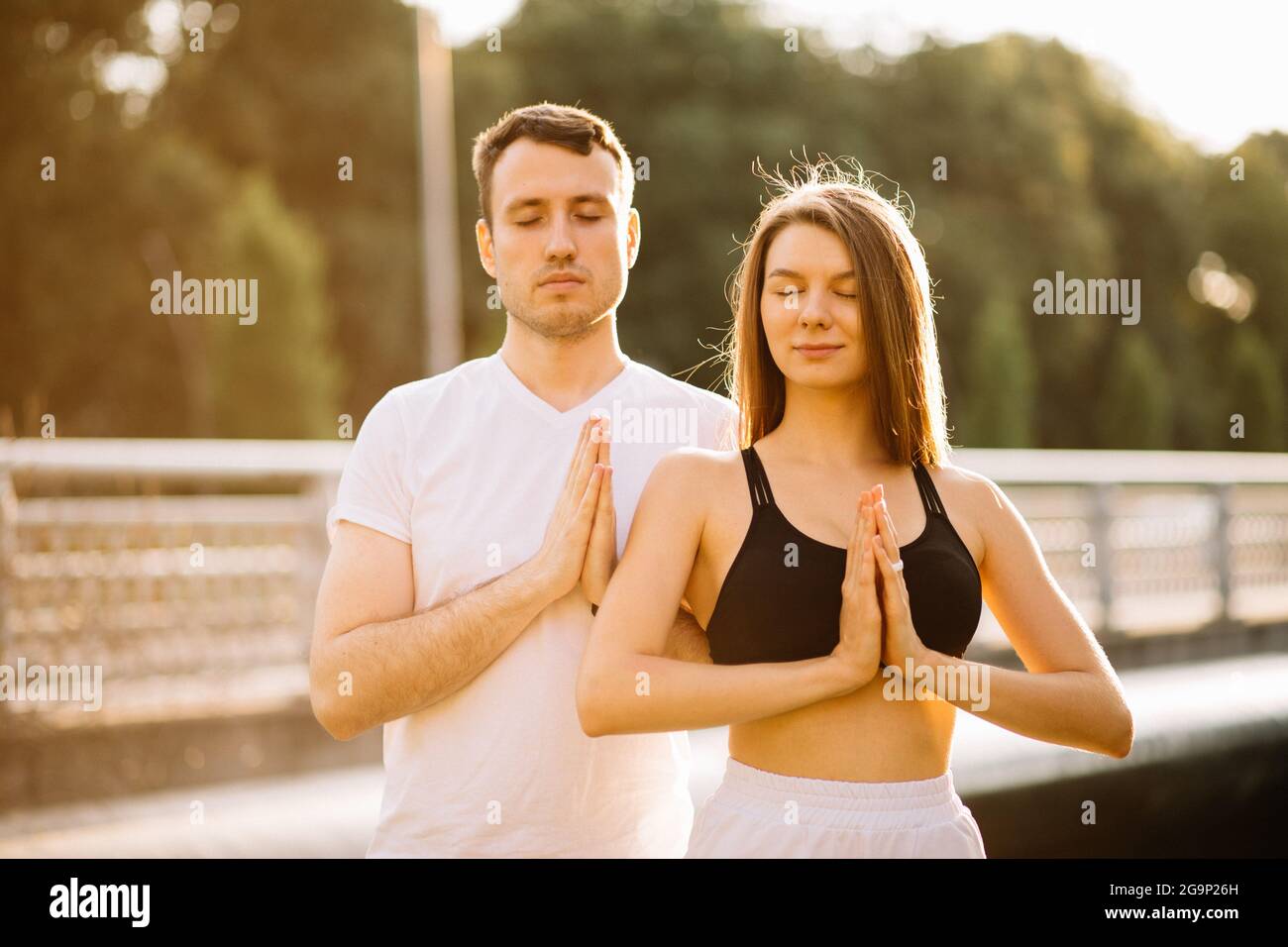 Junge Paar Mann und Frau meditieren gemeinsam beim Sonnenuntergang, während sie stehen, Yoga auf dem Rasen der Stadt, Sommerabend, Lifestyle Stockfoto
