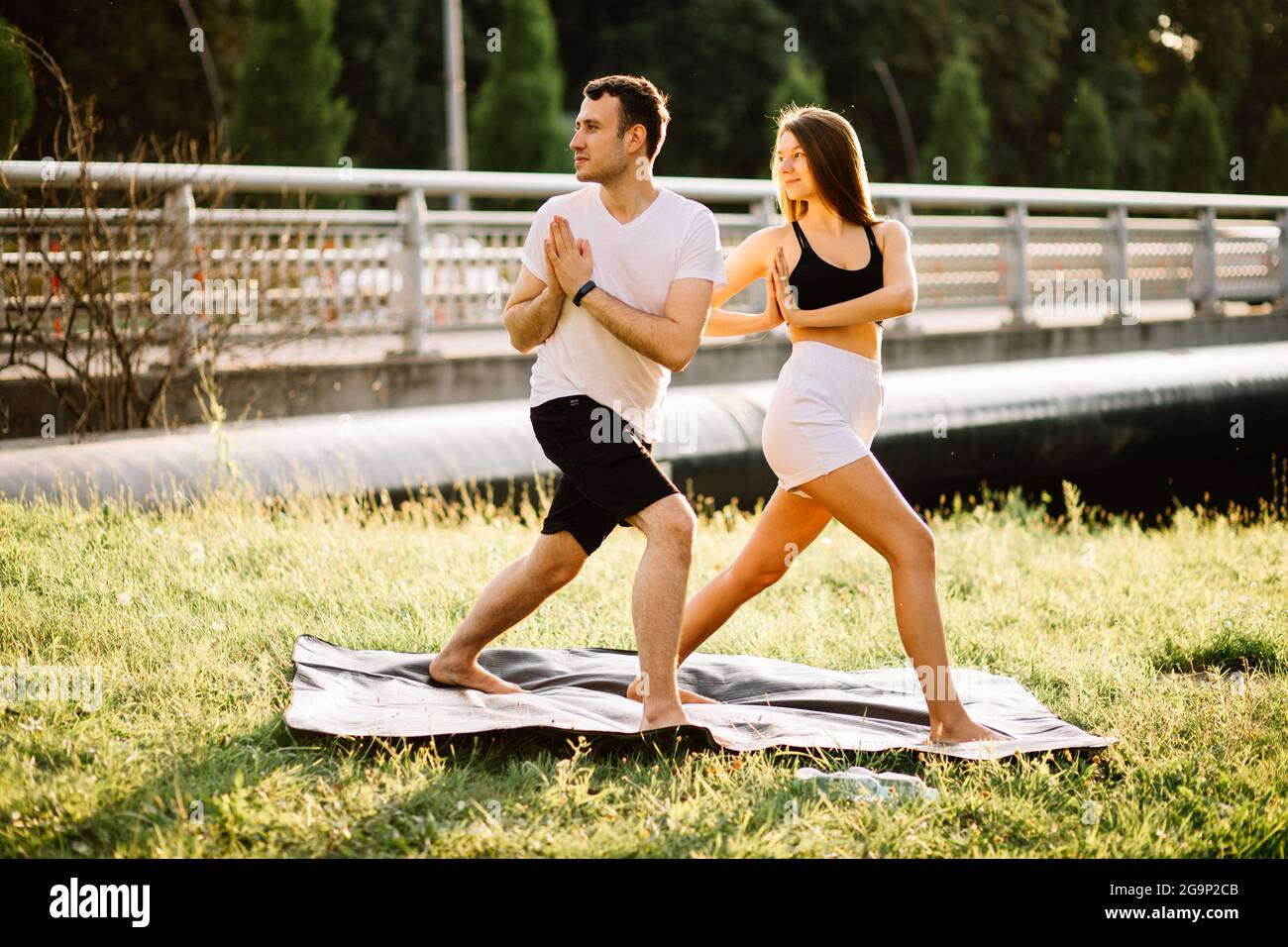 Junges Paar, Mann und Frau, die Sport treiben, Yoga auf dem Rasen der Stadt, Sommerabend, sich bei Sonnenuntergang gemeinsam dehnen, Konzentration auf den Sonnenuntergang Stockfoto