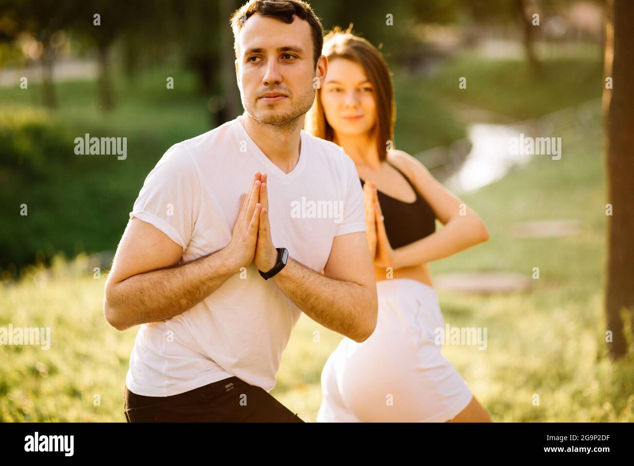 Junges Paar, Mann und Frau, die Sport treiben, Yoga auf dem Rasen der Stadt, Sommerabend, sich bei Sonnenuntergang gemeinsam dehnen, Konzentration auf den Sonnenuntergang Stockfoto