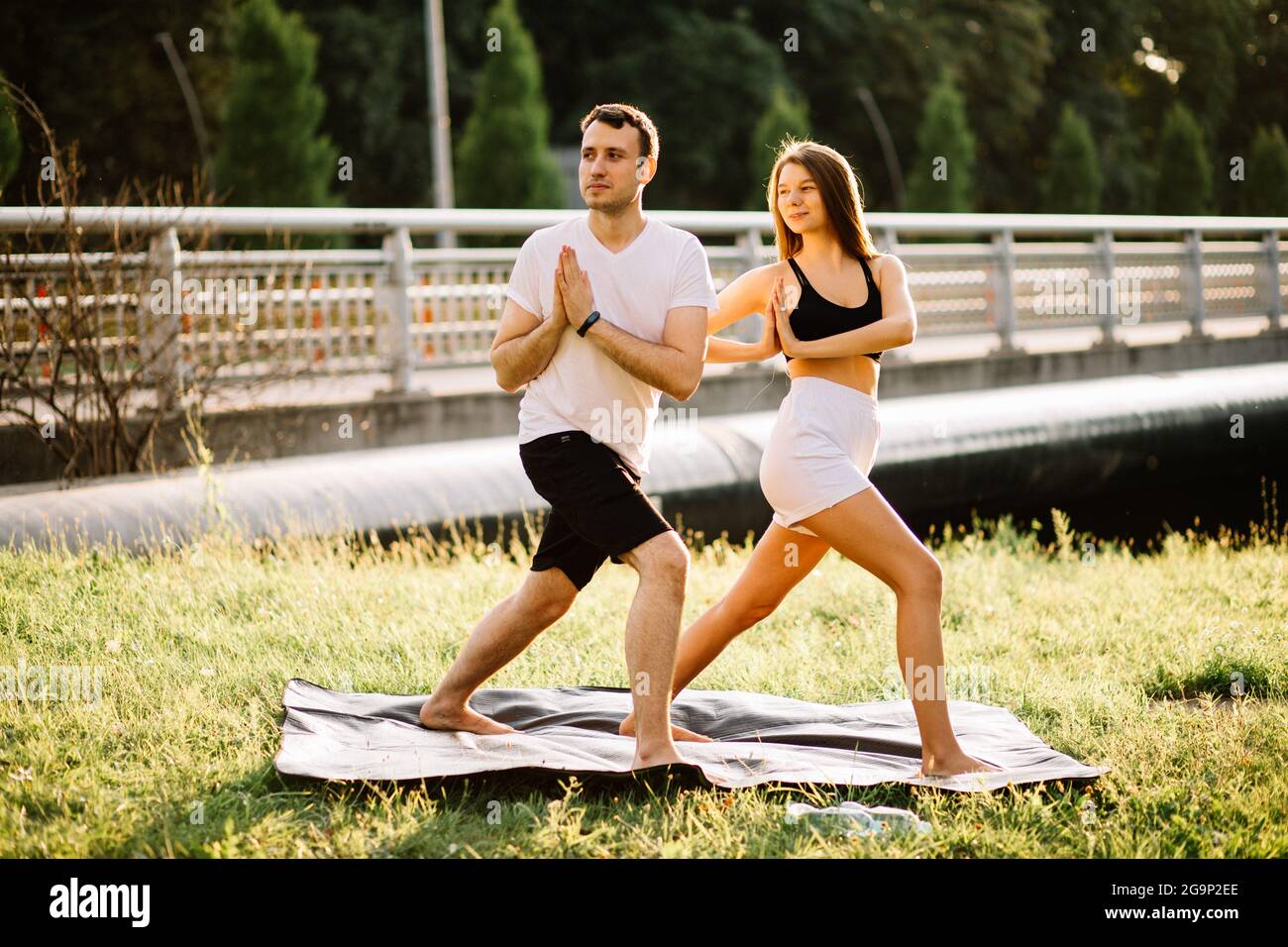 Junges Paar, Mann und Frau, die Sport treiben, Yoga auf dem Rasen der Stadt, Sommerabend, sich bei Sonnenuntergang gemeinsam dehnen, Konzentration Stockfoto