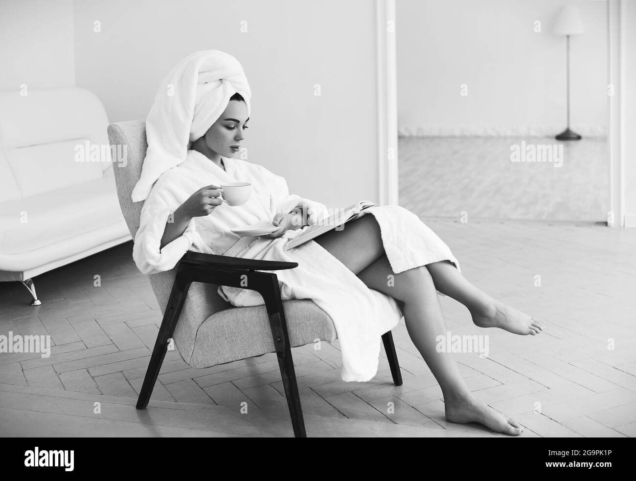 Attraktives Mädchen im Bademantel und Handtuch sitzt auf dem Sessel, trinkt Kaffee und liest Buch. Porträt eines schönen jungen Mädchen zu Hause am Morgen. Schwarz Stockfoto