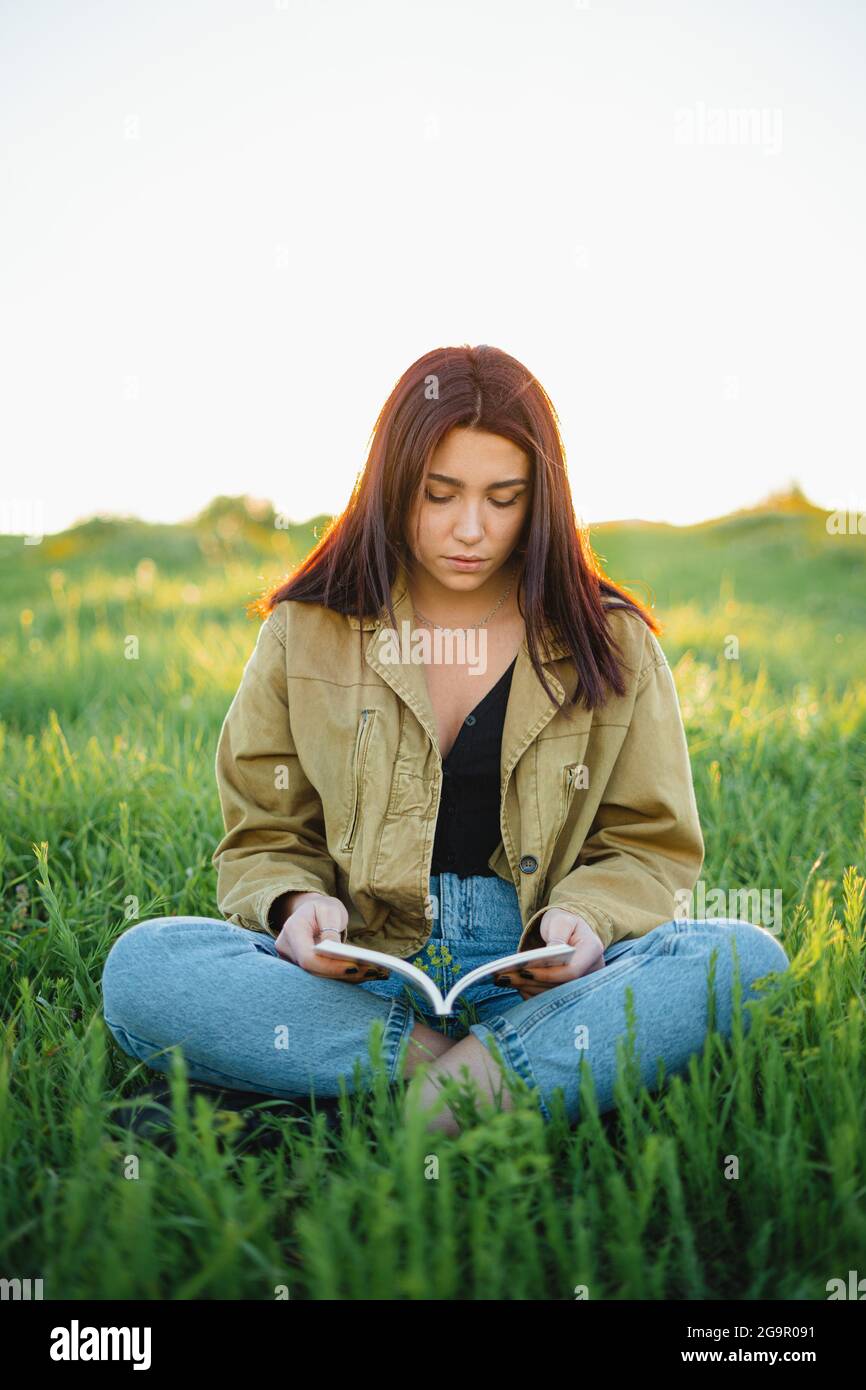 Ein Teenager, der ein Buch bei Sonnenuntergang liest. Sie sitzt im Frühling auf einer grünen Wiese. Stockfoto