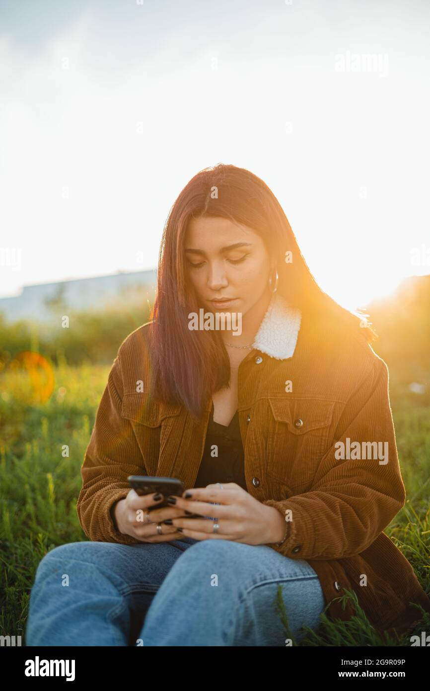 Ein Teenager, der während des Sonnenuntergangs auf einer grünen Wiese sitzt, ihr Telefon benutzt Stockfoto