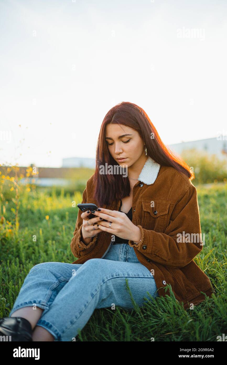 Ein Teenager, der während des Sonnenuntergangs auf einer grünen Wiese sitzt, eine SMS an ihre Freunde am Telefon sendet Stockfoto