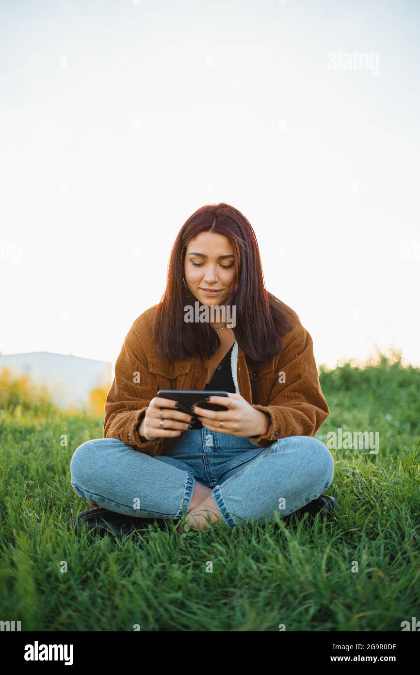 Eine Teenager spielt ein Videospiel auf ihrem Telefon, während sie während des Sonnenuntergangs im Frühling auf einer grünen Wiese sitzt Stockfoto