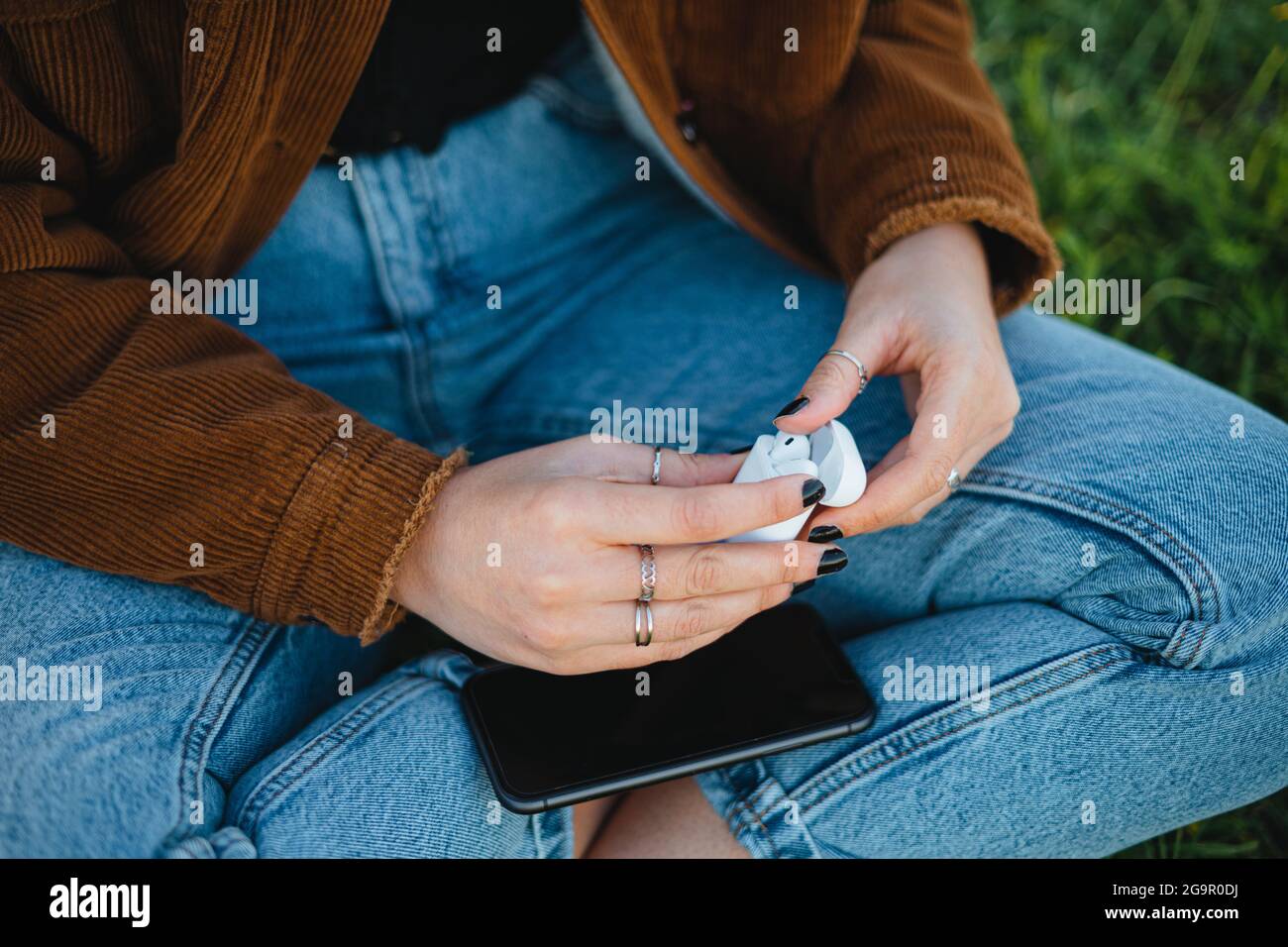 Eine Frau, die die Ladebox mit kabellosen Ohrstöpseln öffnet, während sie auf dem Gras sitzt Stockfoto