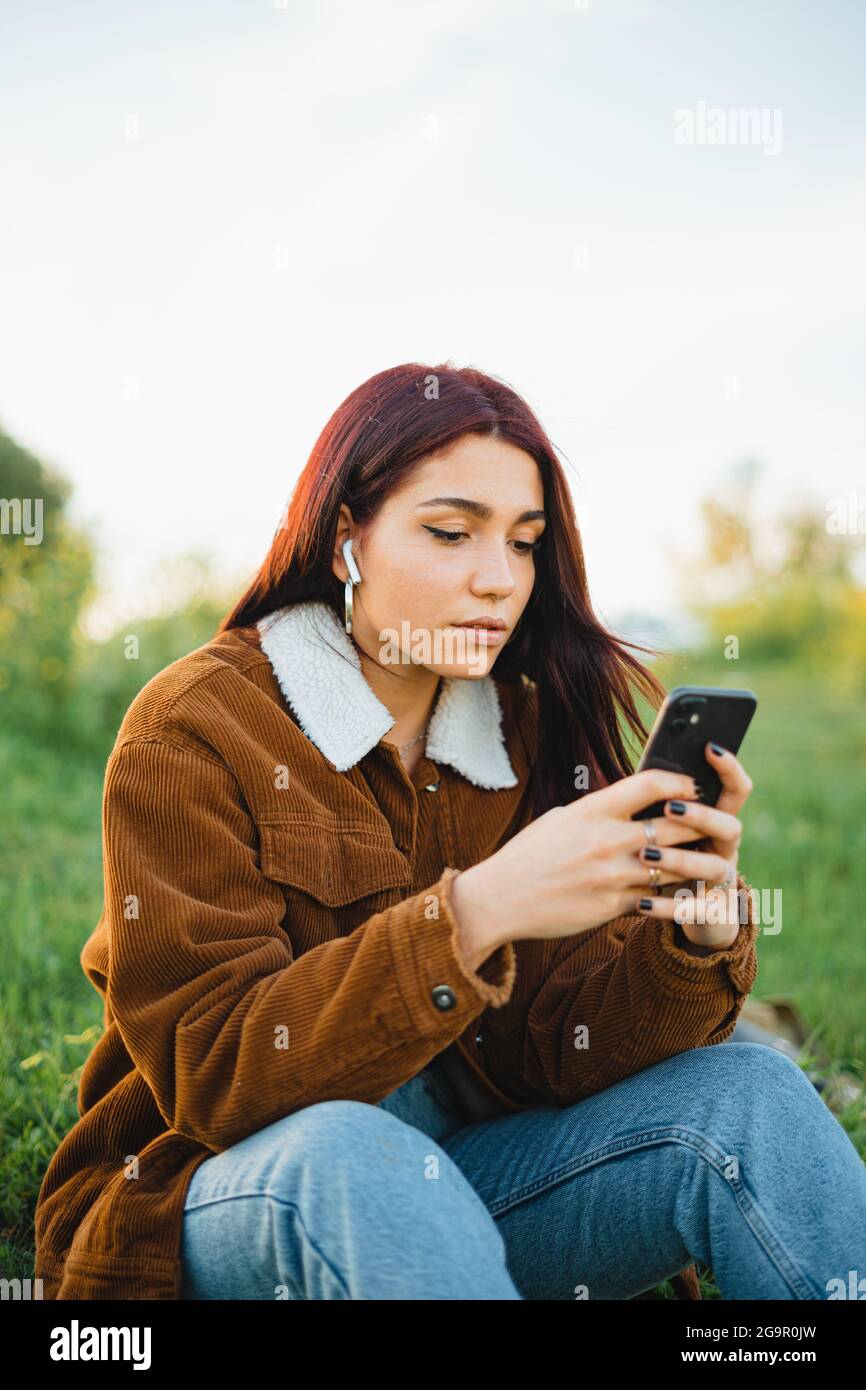 Ein Teenager-Mädchen chattet online mit Freunden am Telefon, während es während des Sonnenuntergangs auf einer grünen Wiese sitzt. Sie hört auch Musik. Stockfoto