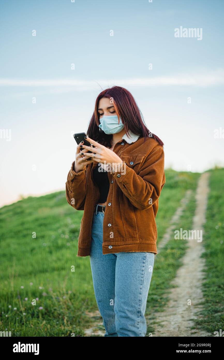 Teenager-Mädchen mit Smartphone auf einem Fußweg auf einem Hügel. Sie trägt eine Gesichtsmaske. Stockfoto