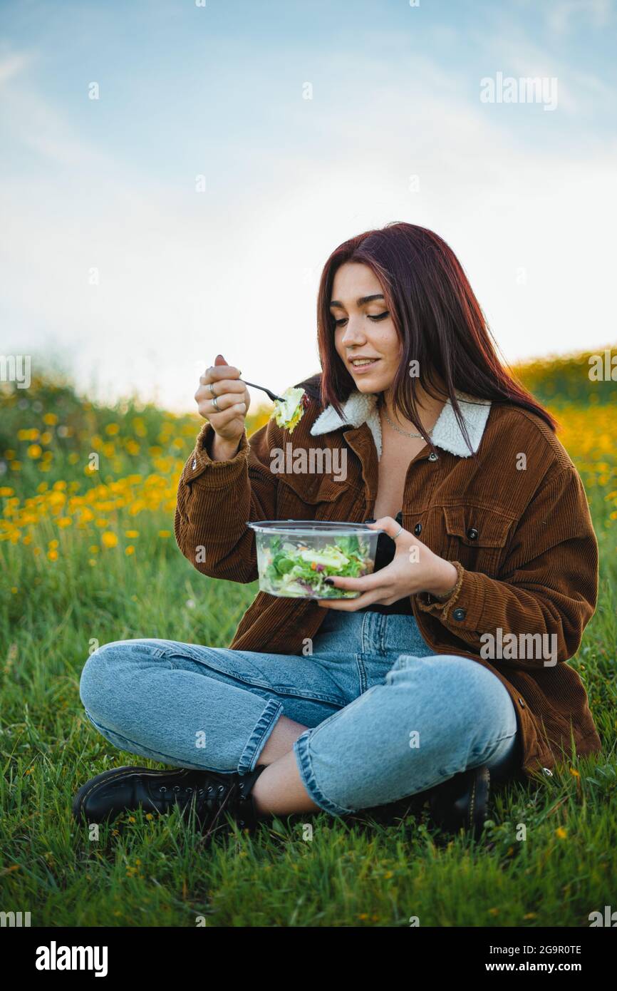 Teenager Mädchen essen einen Salat, während sie auf Gras auf einem Hügel, umgeben von gelben Blumen sitzen. Sie genießt die Ruhe. Stockfoto