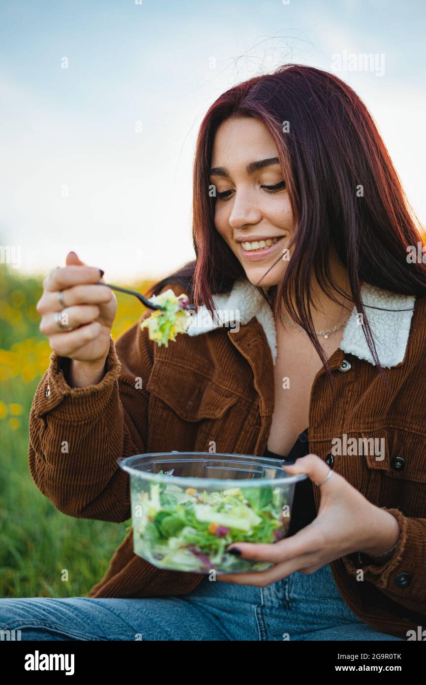 Glückliches Teenager-Mädchen, das einen Salat isst, während es auf einem Hügel auf Gras sitzt. Sie genießt die Ruhe. Stockfoto