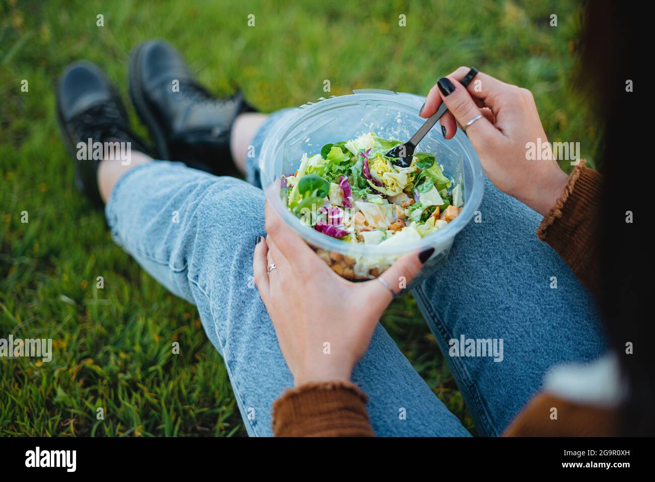 Nahaufnahme einer Salatschüssel auf den Beinen eines Teenagers. Sie sitzt auf Gras und genießt die Natur. Horizontale Aufnahme. Stockfoto