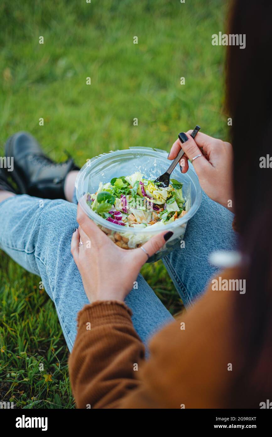Nahaufnahme einer Salatschüssel auf den Beinen eines Teenagers. Sie sitzt auf Gras und genießt die Natur. Vertikale Aufnahme. Stockfoto