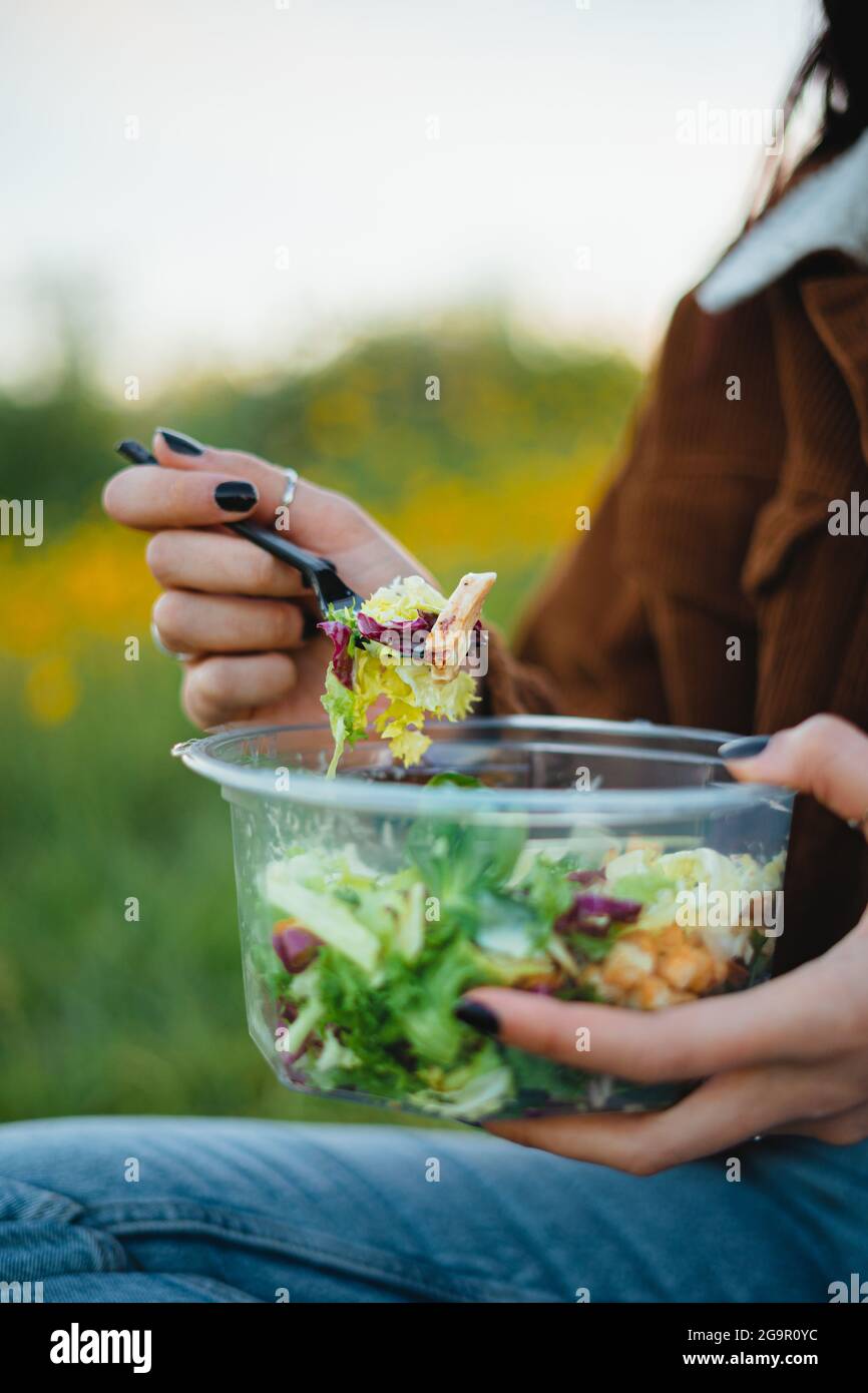 Nahaufnahme einer Salatschüssel. Teenager essen Salat mit schwarzer Kunststoffgabel, während sie auf Gras sitzen, umgeben von gelben Blumen. Stockfoto
