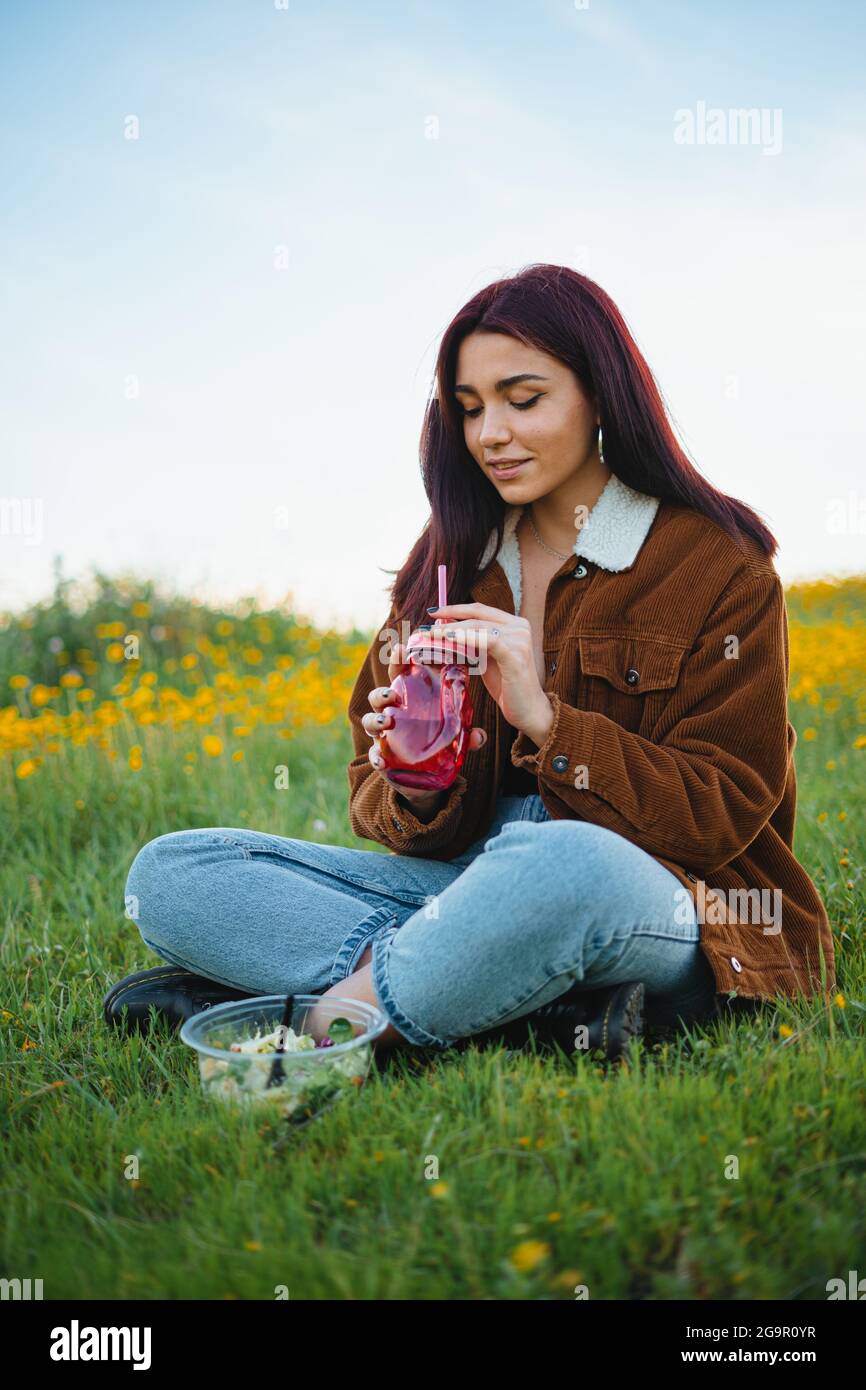 Teenager-Mädchen, das Wasser in einem roten Glas trinkt, das auf dem Gras eines Hügels sitzt. Sie hat einen Salat neben sich. Stockfoto