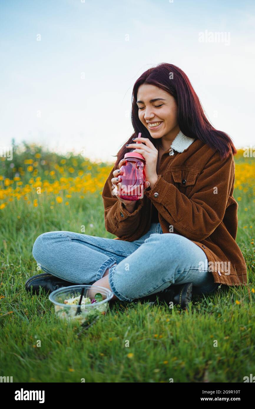 Lachendes Teenager-Mädchen, das Wasser in einem roten Glas trinkt, das auf dem Gras eines Hügels sitzt. Sie hat einen Salat neben sich. Stockfoto