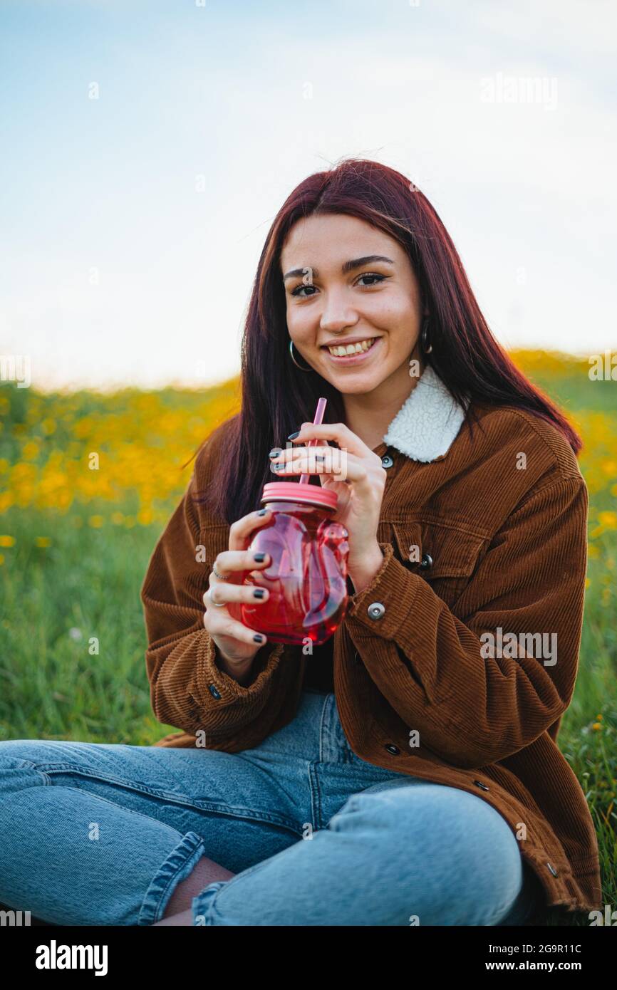 Lächelndes Teenager-Mädchen, das Wasser in einem roten Glas trinkt, das auf dem Gras eines Hügels sitzt. Sie schaut in die Kamera. Stockfoto