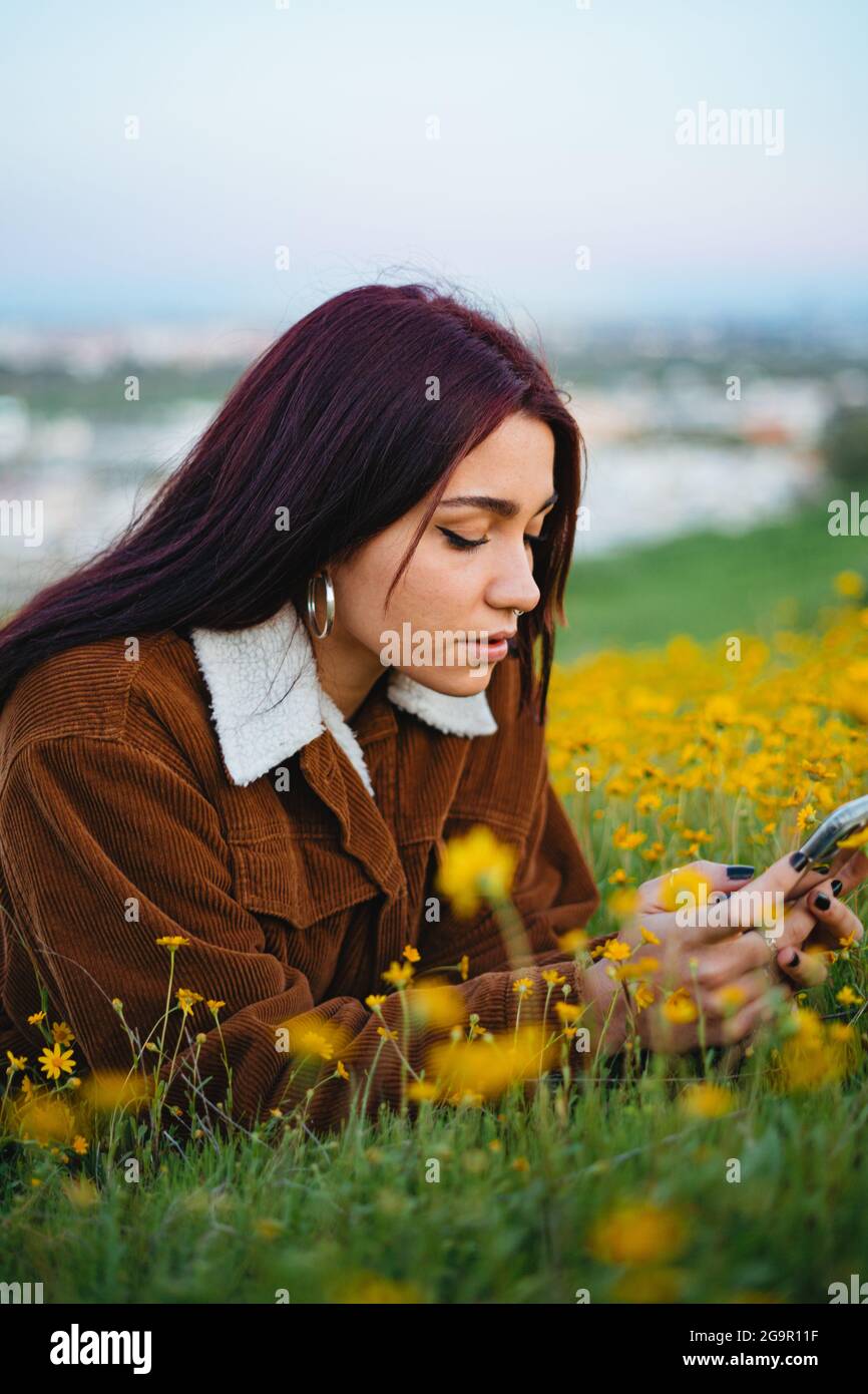 Ein Teenager-Mädchen, das auf ihr Smartphone schaut, während sie sich auf Gras legt. Stadt im Hintergrund. Stockfoto