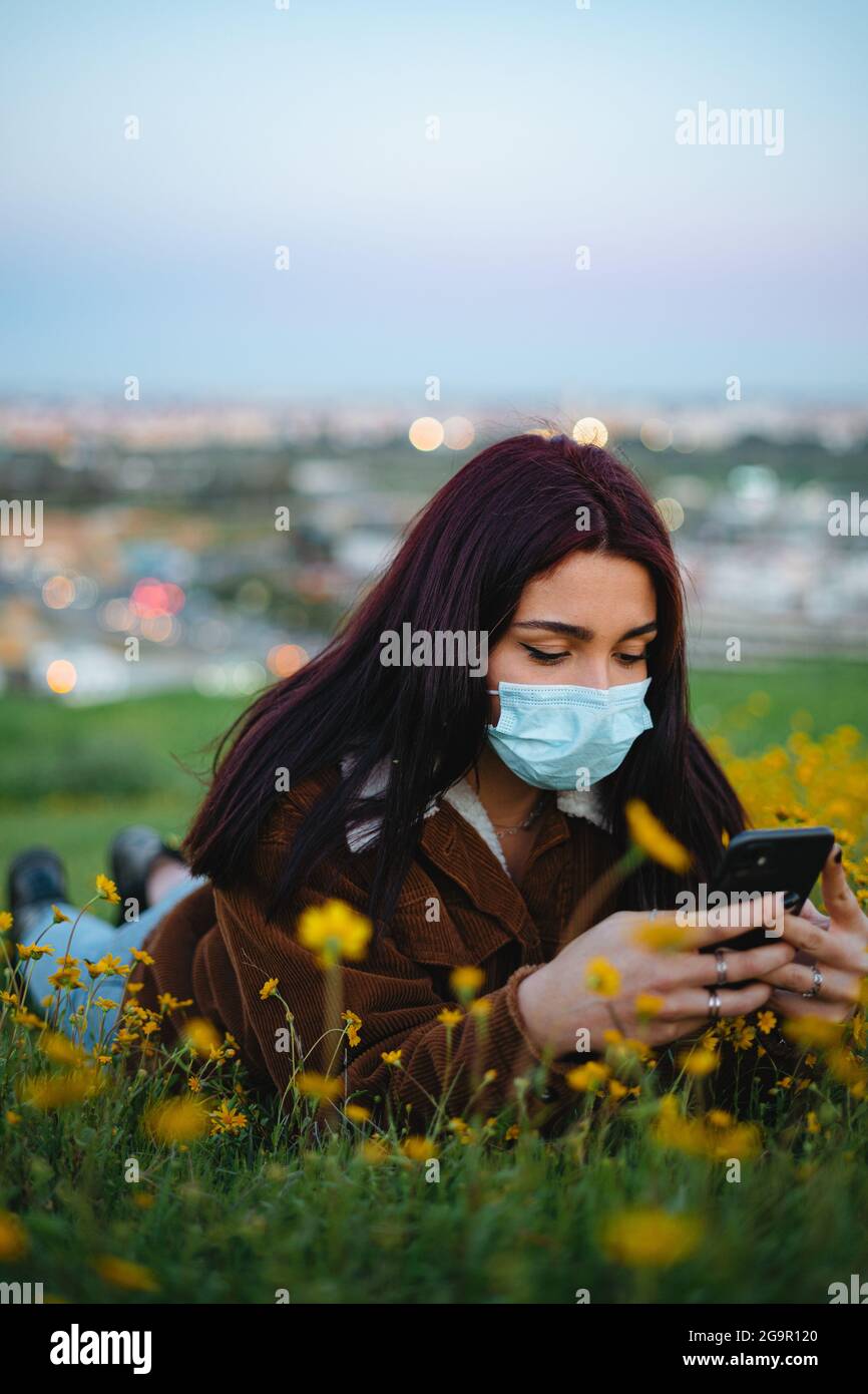 Teenager Mädchen trägt eine medizinische Maske liegt auf Gras. Sie nutzt ihr Smartphone. Stadt im Hintergrund. Stockfoto