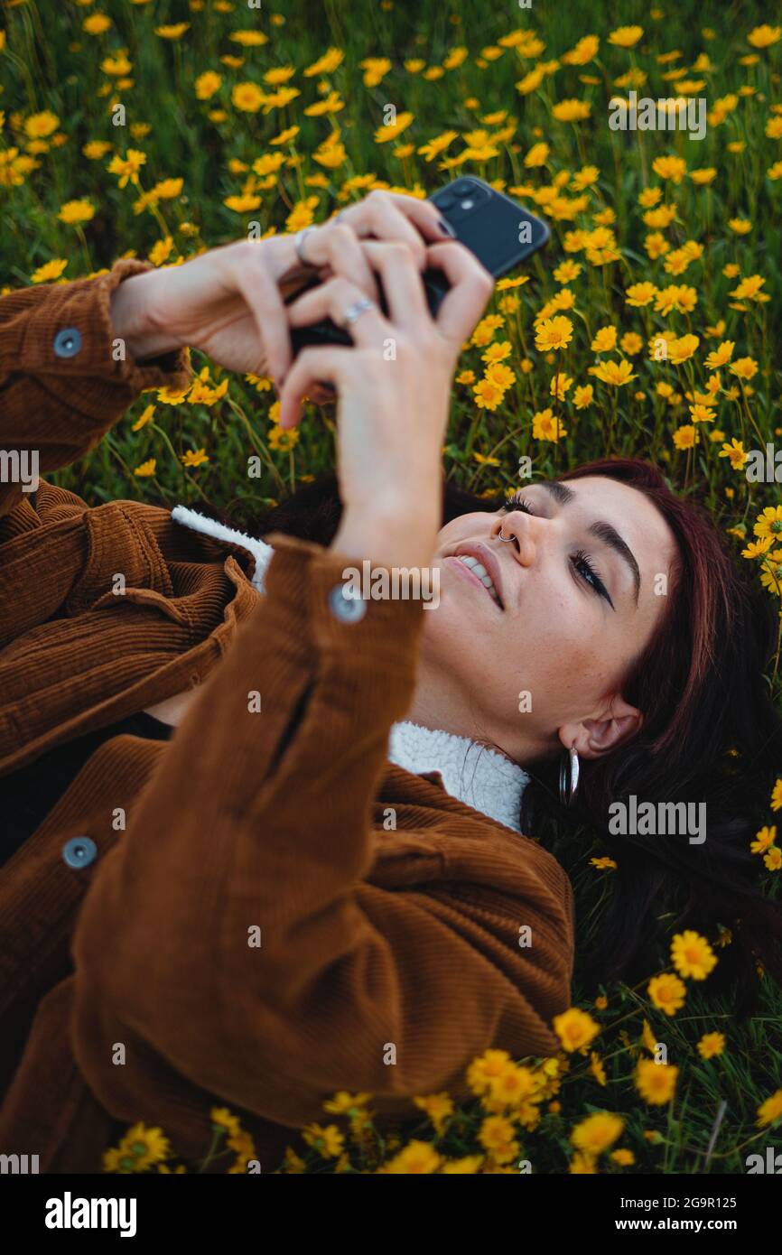 Lächelndes Teenager-Mädchen auf Gras mit Blumen bedeckt entspannen. Sie verwendet das Smartphone, um eine SMS an einen Freund zu senden. Stockfoto