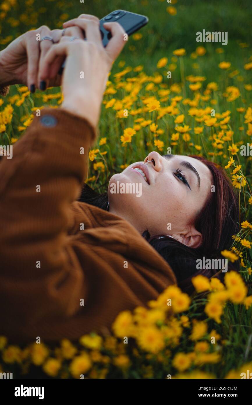 Teenager-Mädchen auf Gras mit Blumen bedeckt entspannen. Sie verwendet das Smartphone, um eine SMS an einen Freund zu senden. (Seitenansicht) Stockfoto