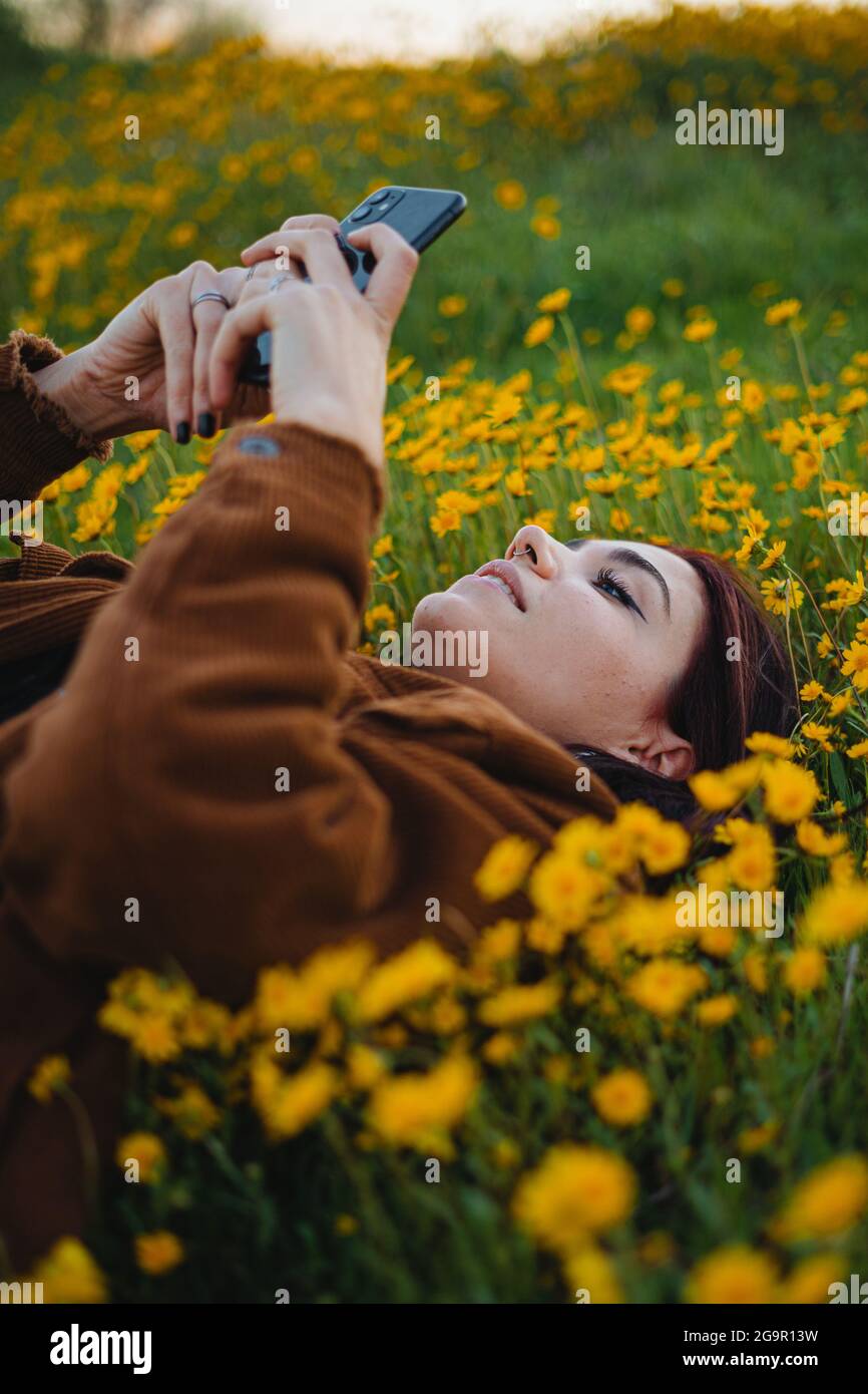 Teenager-Mädchen entspannen während des Sonnenuntergangs auf Gras mit Blumen bedeckt. Sie nutzt das Smartphone, um Spiele zu spielen. Stockfoto