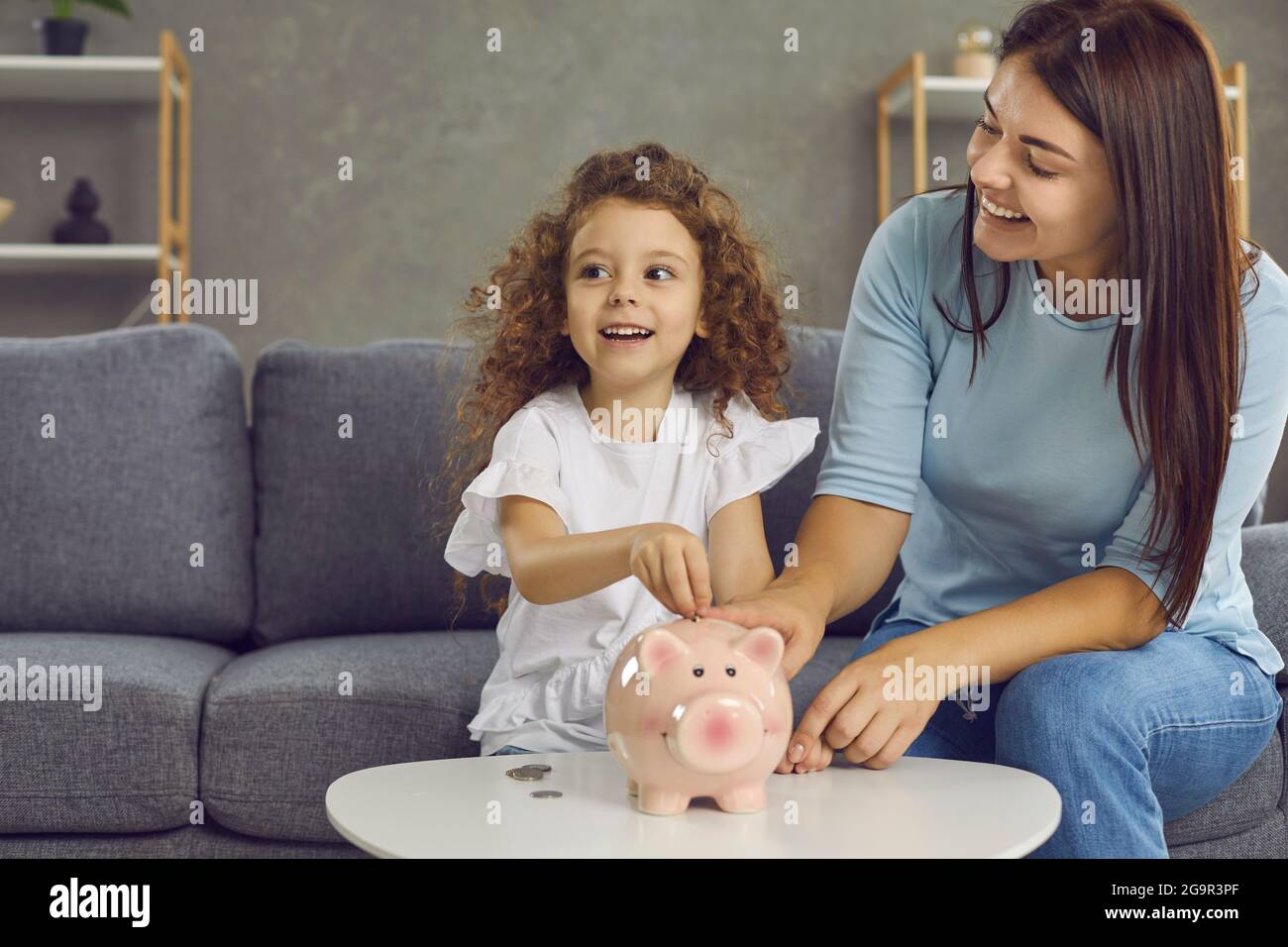 Glückliche junge Mutter und kleine Tochter, die zusammen Münzen in ihre Sparschwein legen Stockfoto