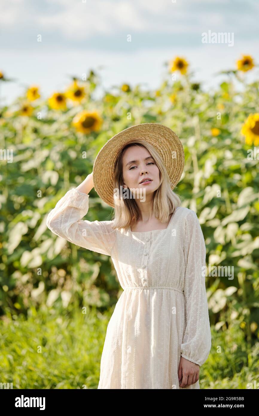 Glückliche junge blonde Frau in Hut und weißen romantischen Kleid Stehen auf grünem Gras vor der Kamera gegen Sonnenblume Feld an sonnigen Tag Stockfoto