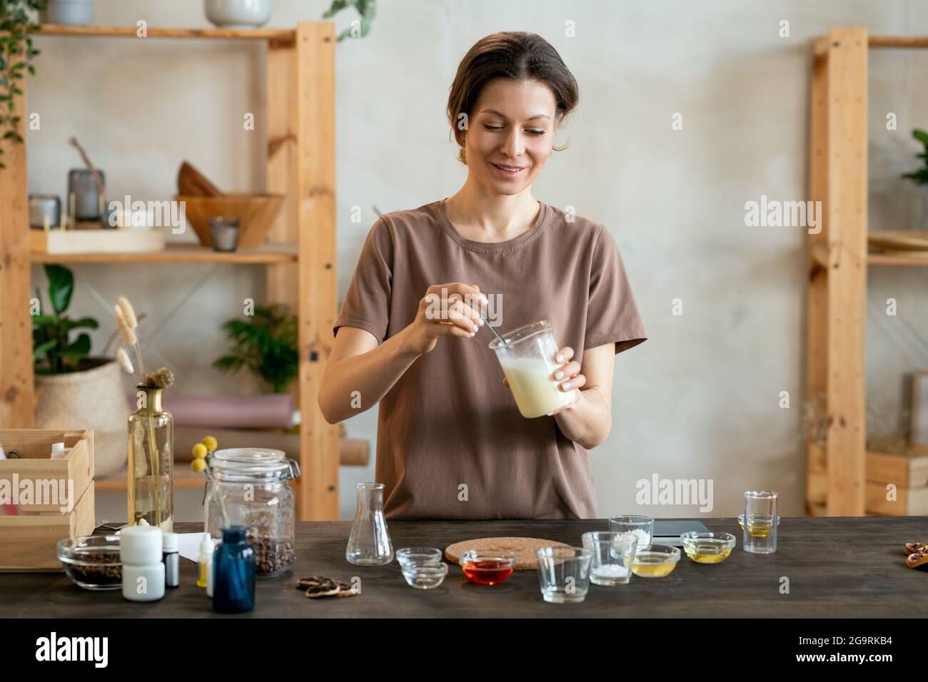 Glückliche junge Frau hält Glaswaren mit geschmolzener Seifenmasse über Tisch bei der Herstellung Mischung für natürliche handgemachte kosmetische Produkte Stockfoto