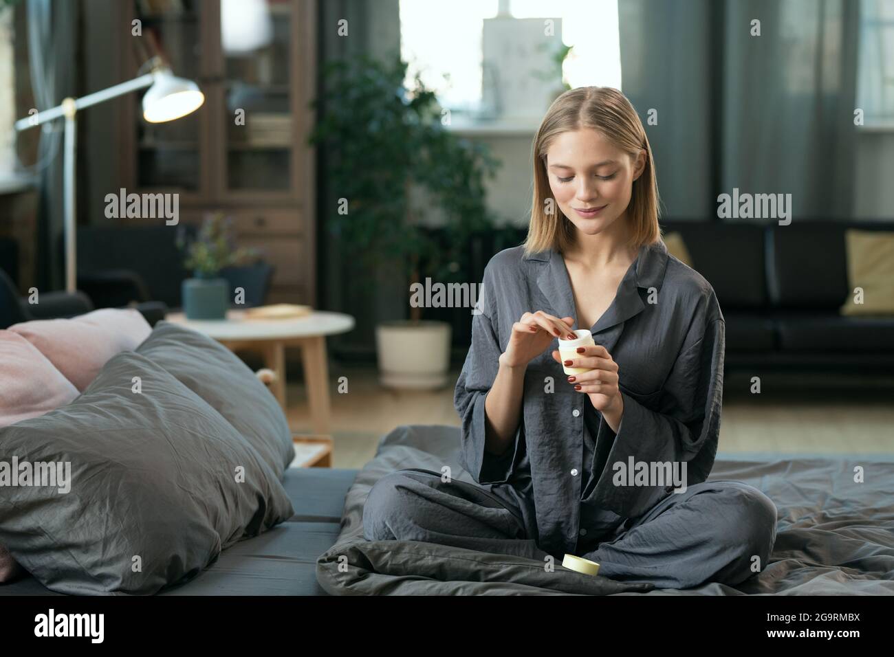 Junge blonde hübsche weibliche in grauen Pyjamas Anwendung natürliche Handarbeit Kosmetisches Produkt auf ihrem Gesicht, während sie auf dem Bett innen sitzt Am Morgen Stockfoto