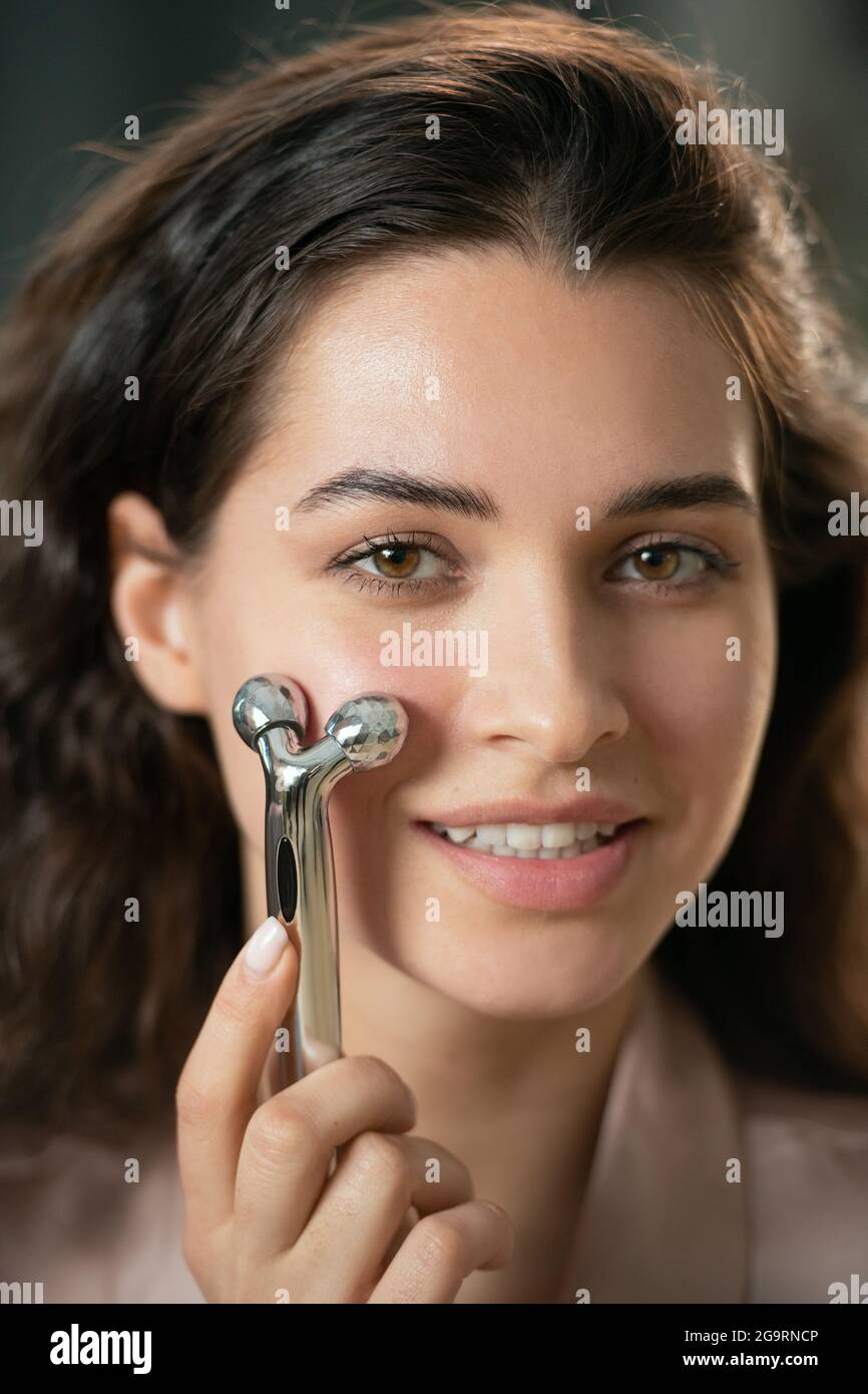 Wunderschöne junge Brünette Frau mit metallischen Massage Roller auf sie Gesicht und Blick auf Sie mit einem Lächeln, während Schönheit genießen Verfahren Stockfoto
