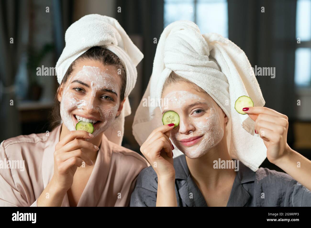 Junge Frauen in Seiden-Pyjamas, Handtücher auf den Köpfen, Tonmasken auf Gesichtern und Gurkenscheiben auf den Augen Spaß haben und genießen Schönheitsverfahren Stockfoto