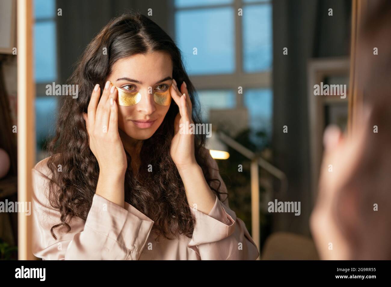 Junge Brünette Frau in Seide Pyjamas setzen goldene Kosmetik unter dem Auge Patches beim Stehen vor dem Spiegel und beim Anschauen Selbst Stockfoto