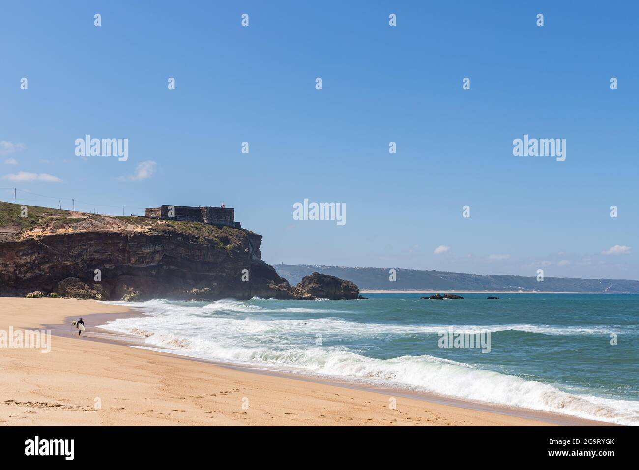 Nazare, Portugal - 29. Juni 2021: Ein Surfer am North Beach geht in Richtung Meer unterhalb der Festung und des Leuchtturms von Nazare Stockfoto