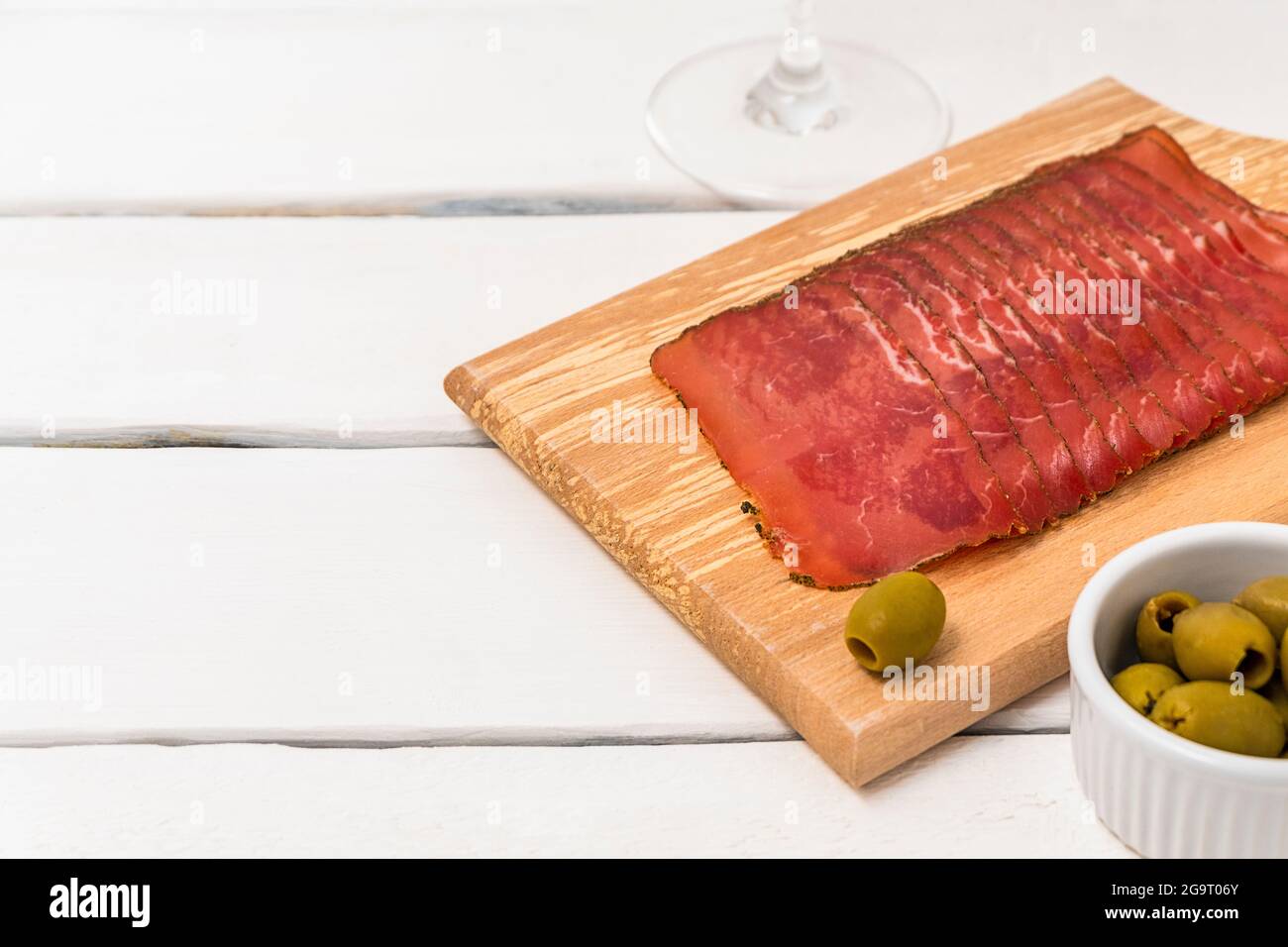 In Scheiben geschnittenes Jamon auf Schneidebrett auf weißem Holztisch mit Oliven, Gewürzen und Kopierplatz Stockfoto