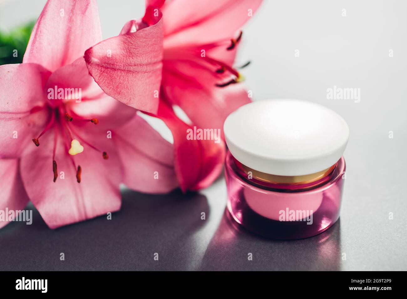 Glas mit Feuchtigkeitscreme, umgeben von rosa Lilienblumen auf grauem Hintergrund. Bio-Kosmetik. Hautpflege Stockfoto