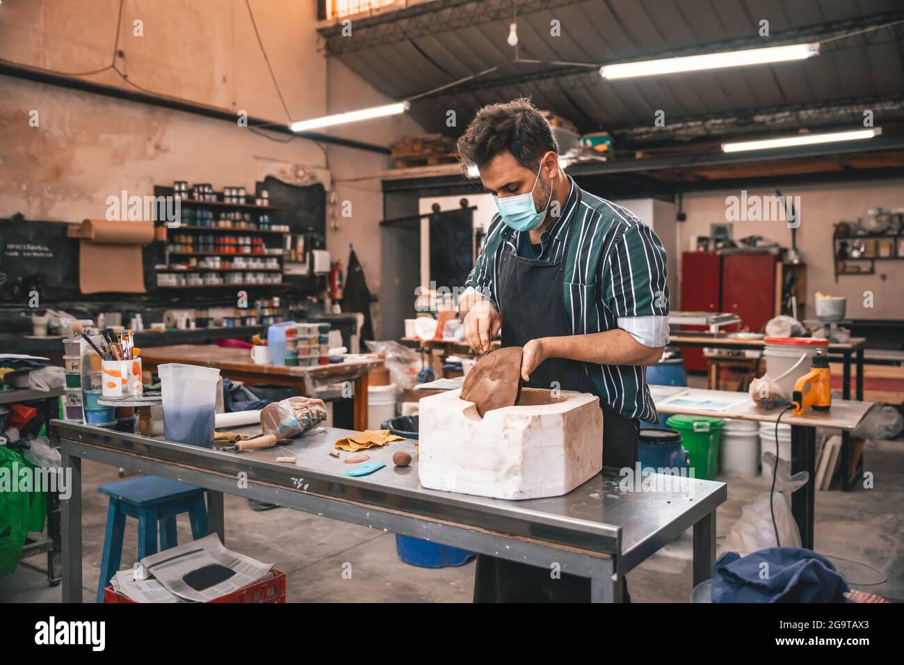 Ein erwachsener hispanischer Mann trägt eine Gesichtsmask und macht in einer Werkstatt einen Tontopf auf einem Tisch Stockfoto