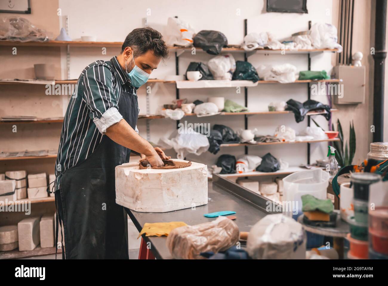 Ein erwachsener hispanischer Mann trägt eine Gesichtsmask und macht in einer Werkstatt einen Tontopf auf einem Tisch Stockfoto