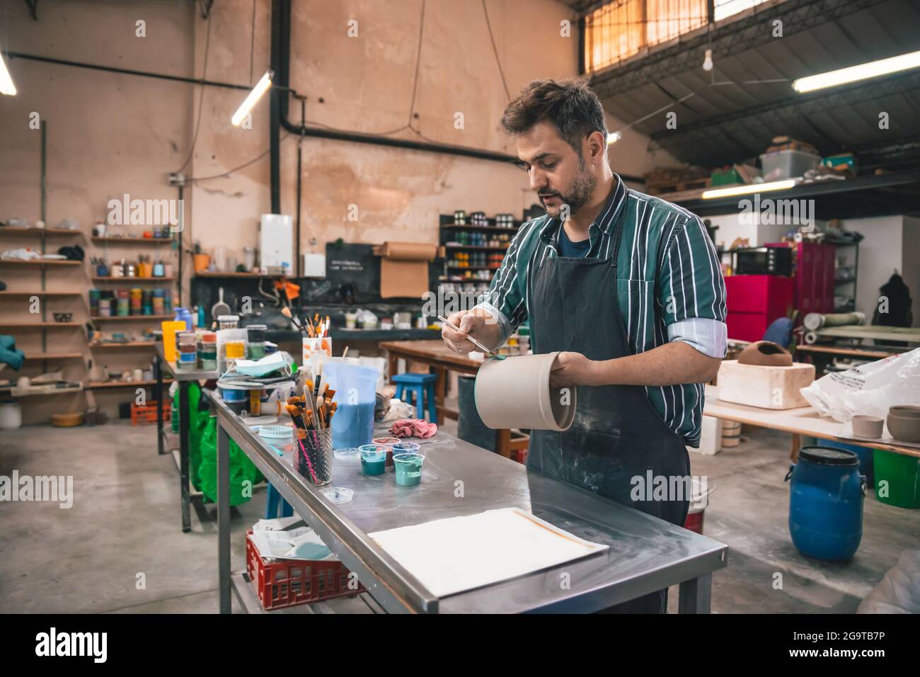 Ein erwachsener hispanischer Mann malte in einer Werkstatt mit verschwommenem Hintergrund einen Tontopf Stockfoto